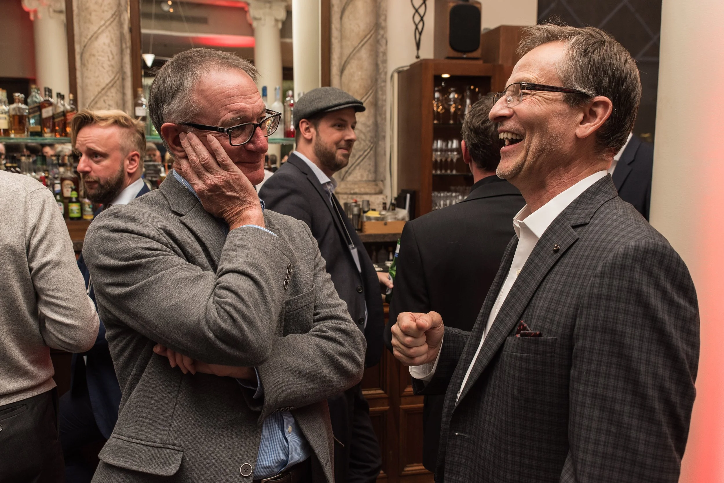 Two middle-aged men in suits smiling and chatting at a social event in a bar or lounge, with other people in the background.