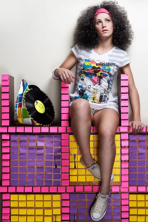 A young woman with curly hair, pink headband, and casual clothing sitting on a colorful, block-like structure with a vinyl record and purse beside her.