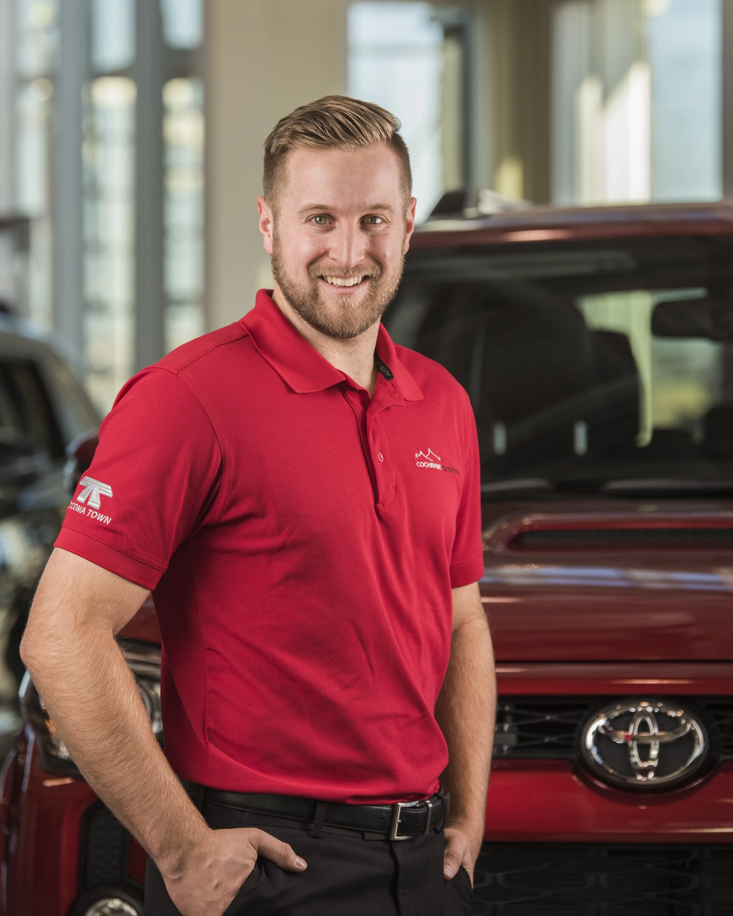 A smiling man with a beard and short hair, wearing a red polo shirt with a logo, standing in a car dealership showroom next to a red Toyota vehicle.