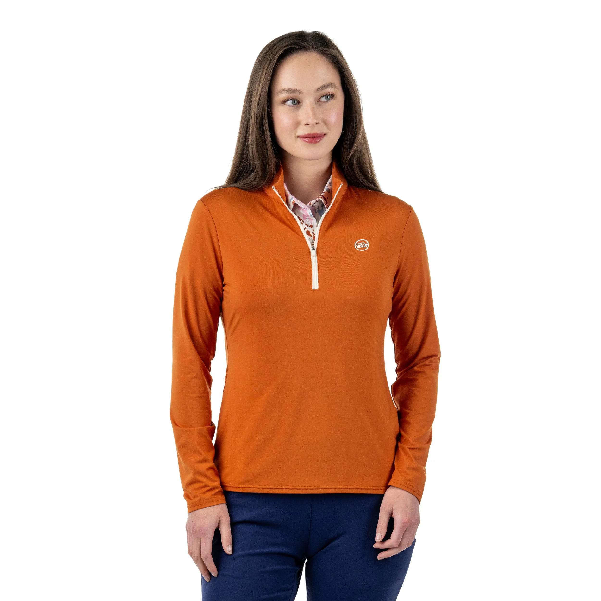 A woman with long brown hair wearing an orange long-sleeve athletic top with a half-zip collar and a small logo on the chest, paired with dark blue pants, standing against a white background.