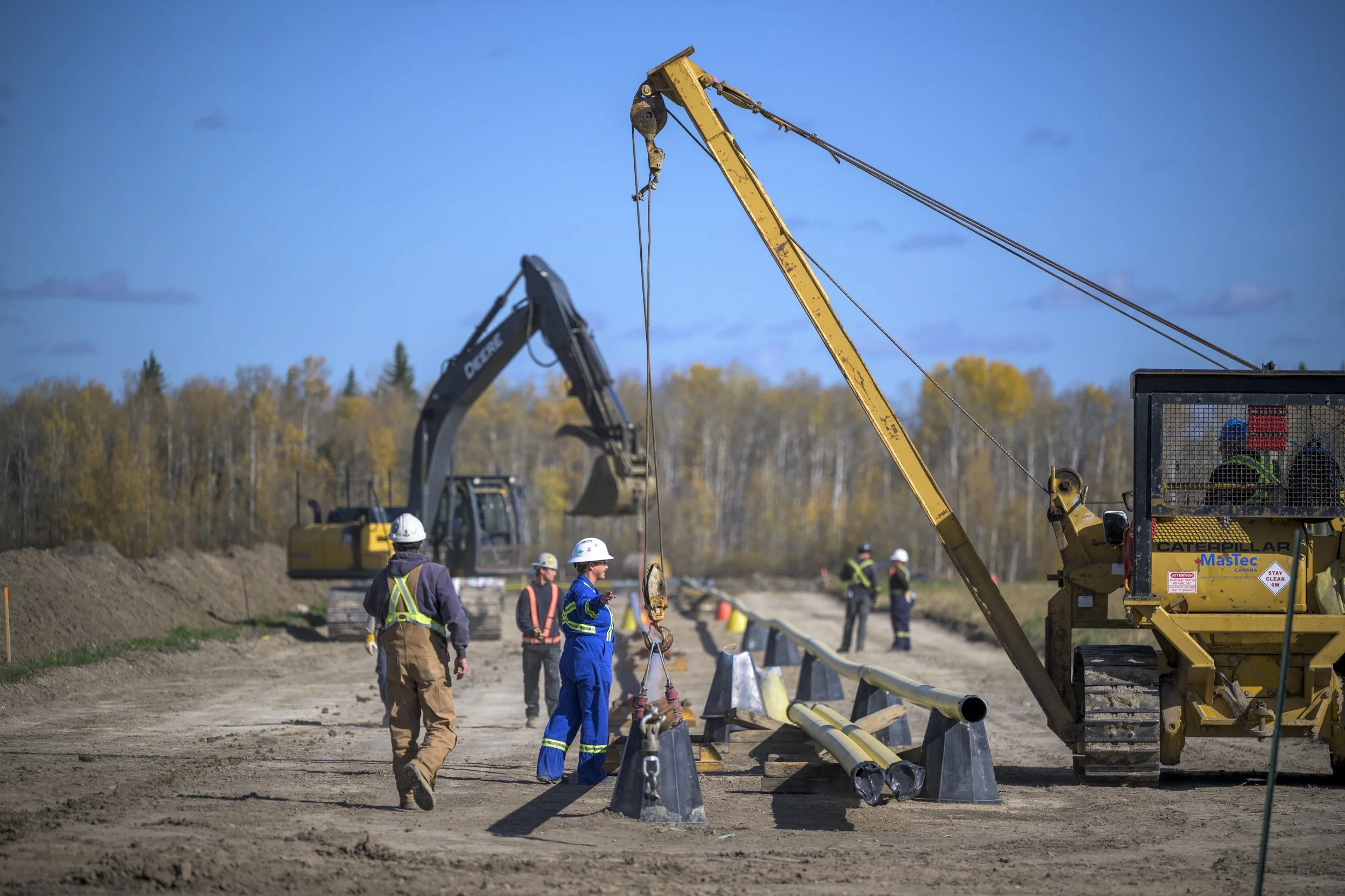 Construction workers on a dirt road using heavy machinery like excavators and cranes, with pipes and safety gear, set against a background of trees and a blue sky.