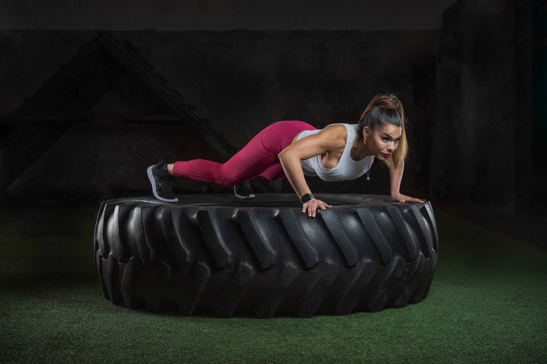 A woman in athletic clothing performing a push-up on a large tractor tire in a gym.