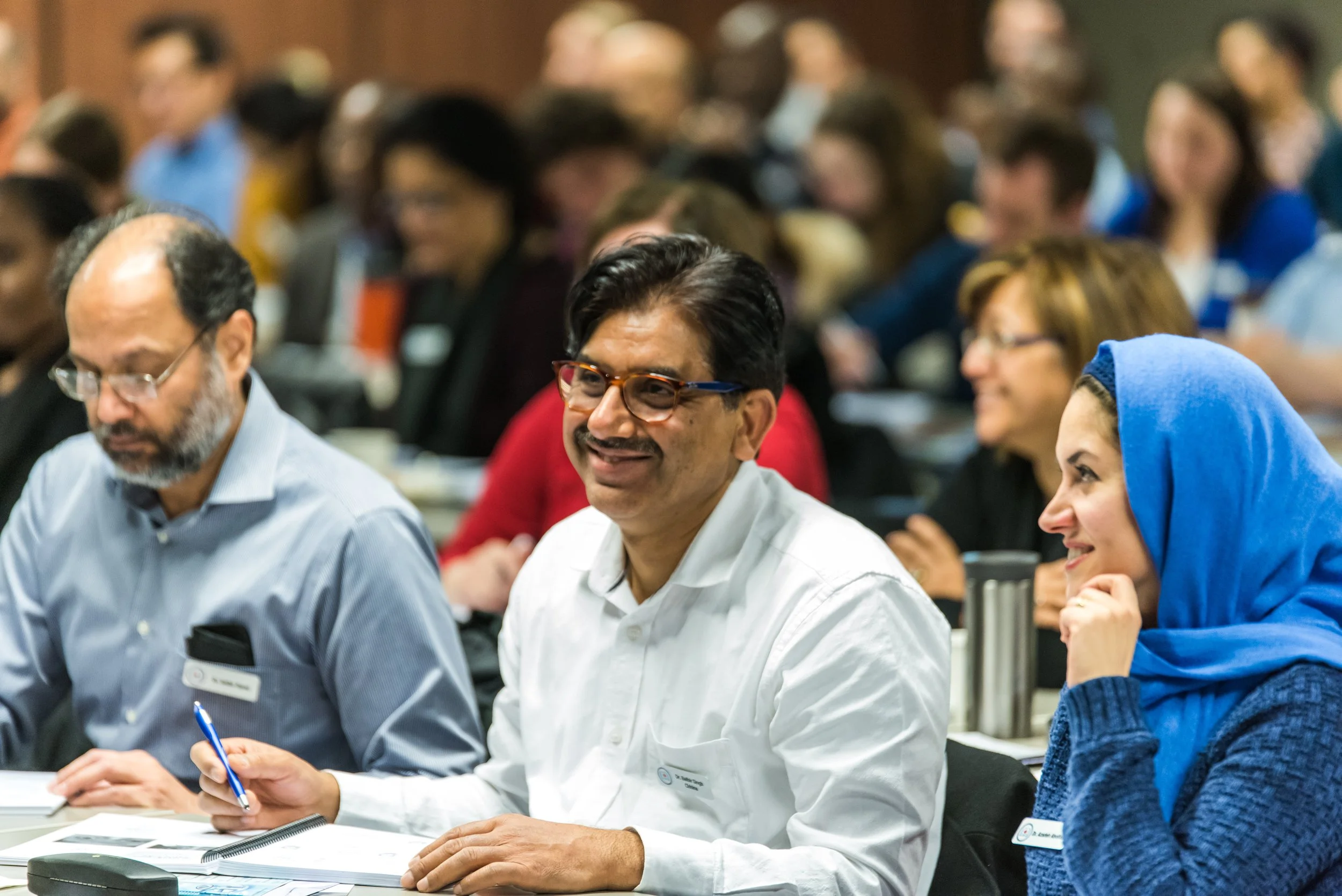 A diverse group of people attending a conference, sitting at tables, with some taking notes and others listening attentively.