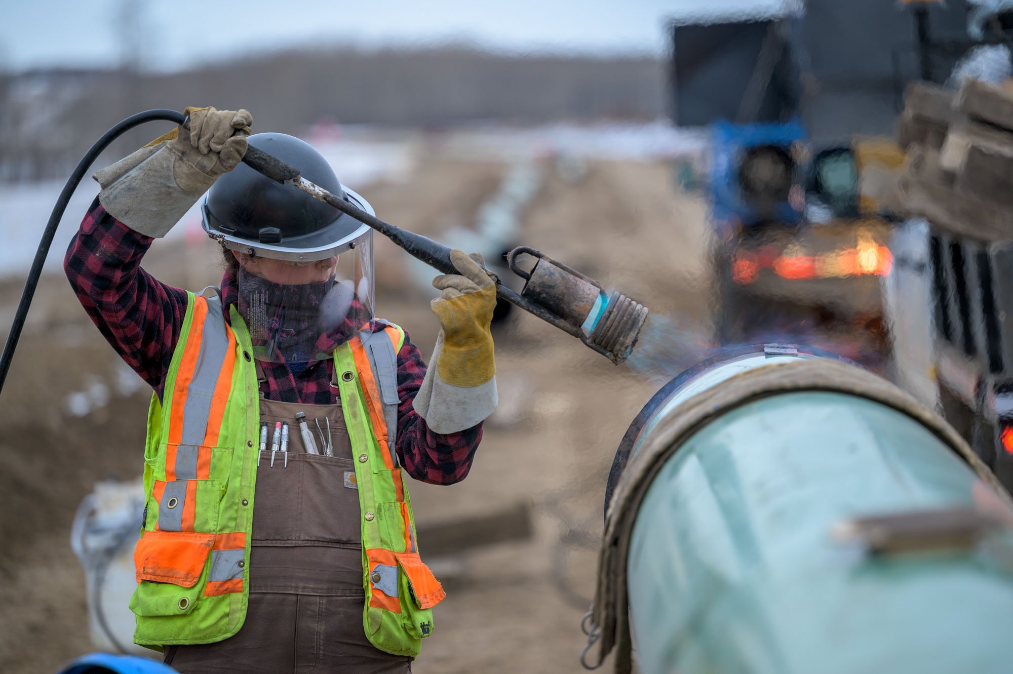 A construction worker wearing a protective helmet, face shield, gloves, and a neon safety vest is using a blowtorch on a large pipe at a construction site.