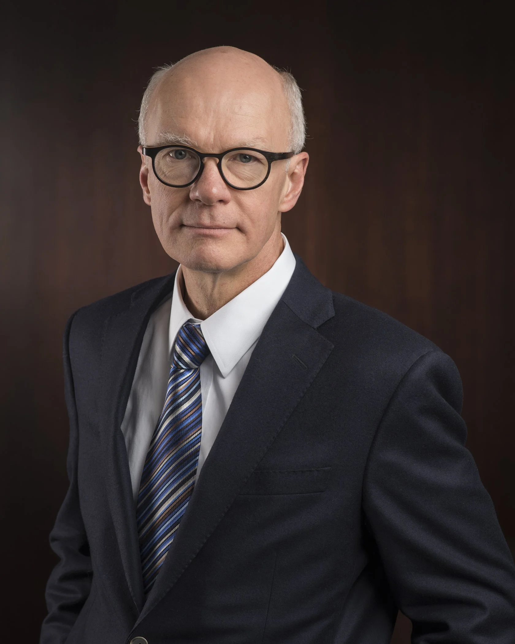 Portrait of an older man with glasses wearing a dark suit, white shirt, and striped tie, against a dark background.