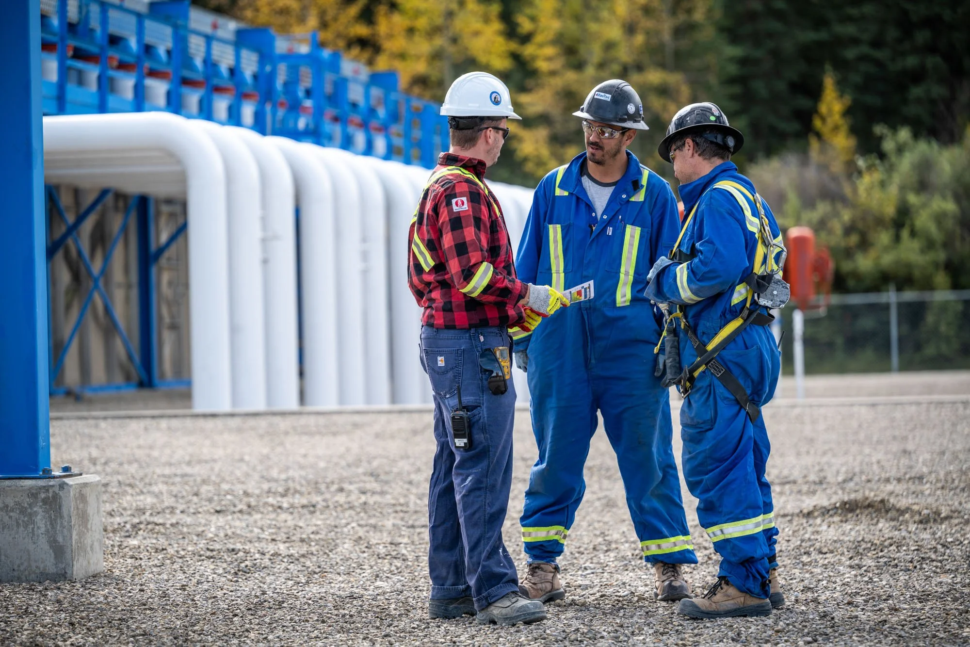 Three workers in safety gear and helmets standing and discussing outdoors near industrial pipes and equipment.