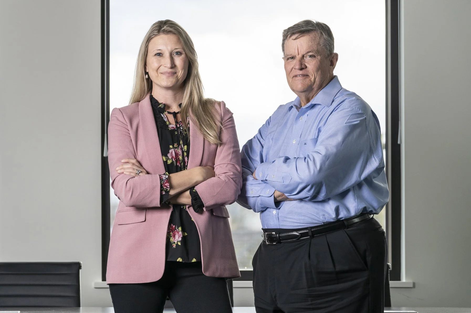 A woman and a man standing with arms crossed in a modern office, smiling, with a large window behind them.