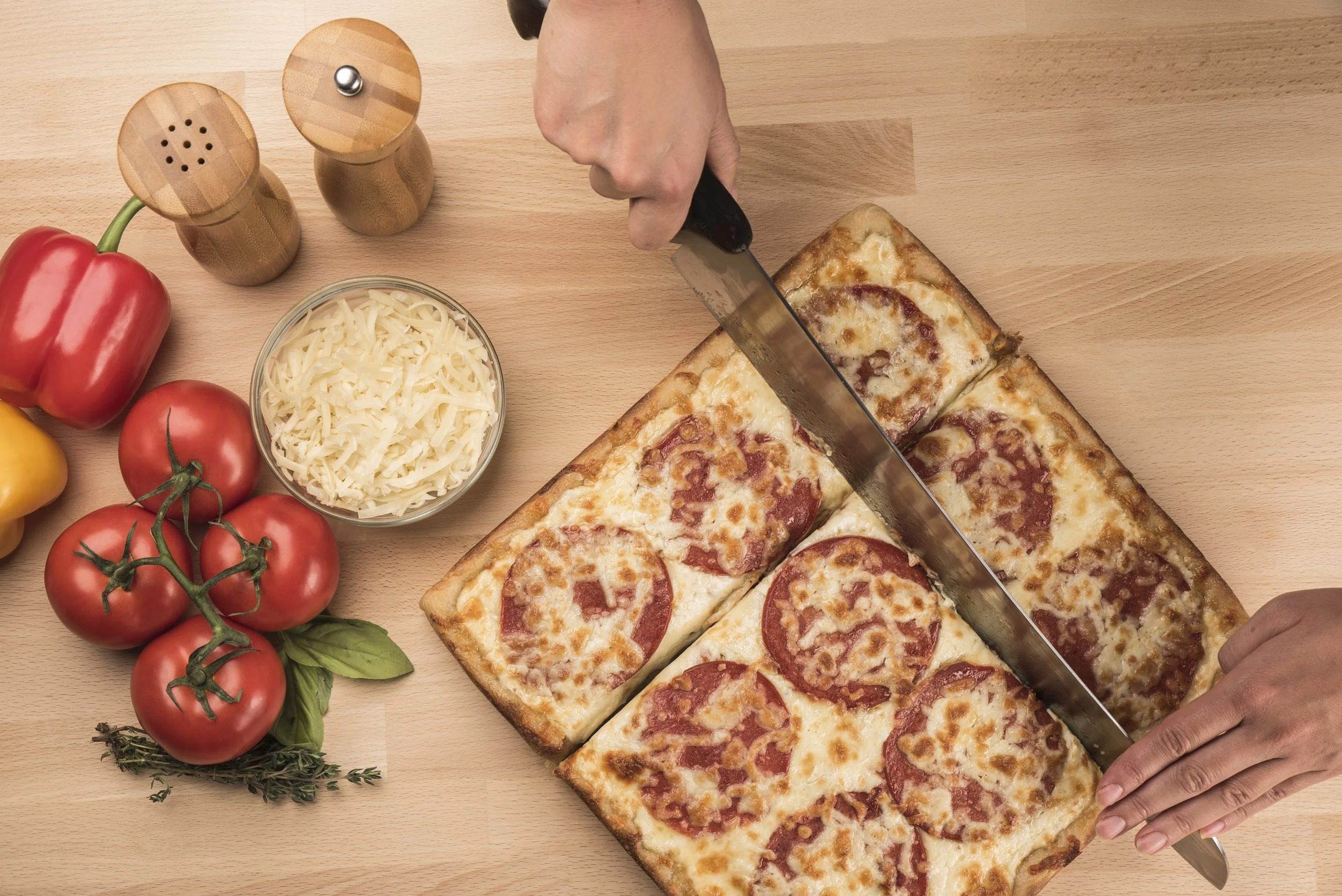 Person slicing a large pepperoni pizza on a wooden table with fresh tomatoes, shredded cheese, red and yellow bell peppers, and spice shakers nearby.