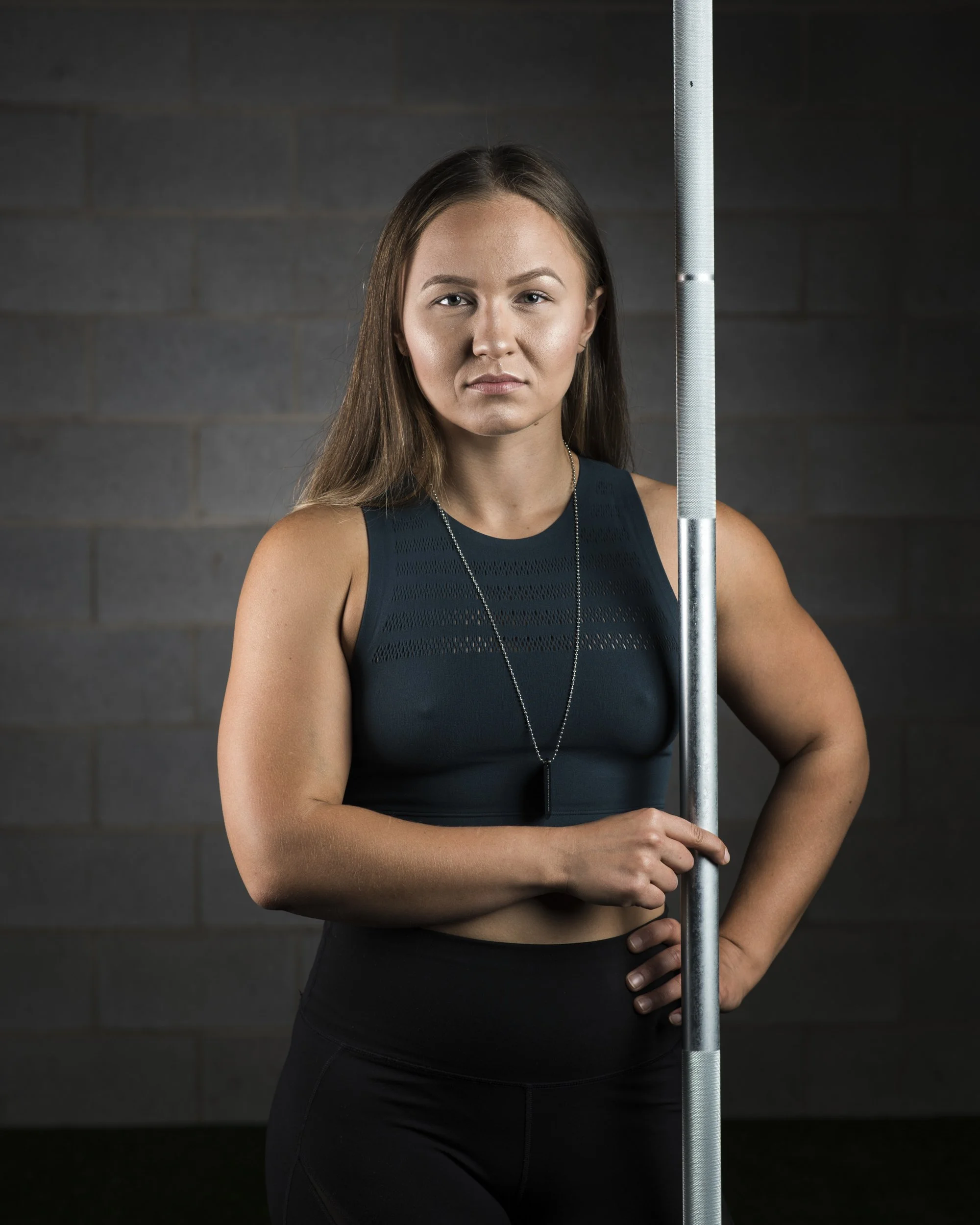A young woman in athletic clothing holding a pole, standing against a dark brick wall.