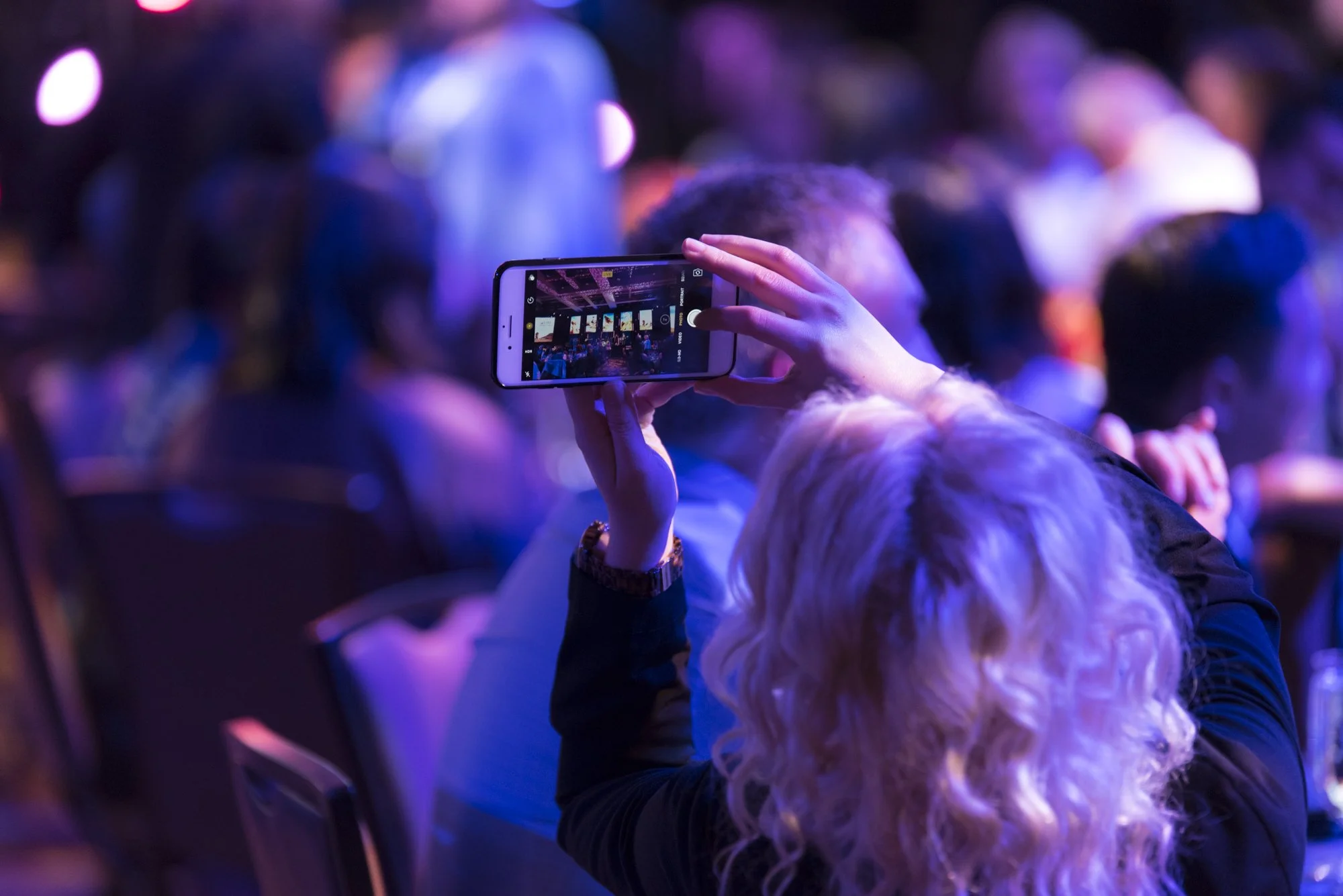 A woman with wavy blonde hair taking a photo or recording a video with her smartphone at a crowded event or concert, illuminated by purple and blue stage lighting.