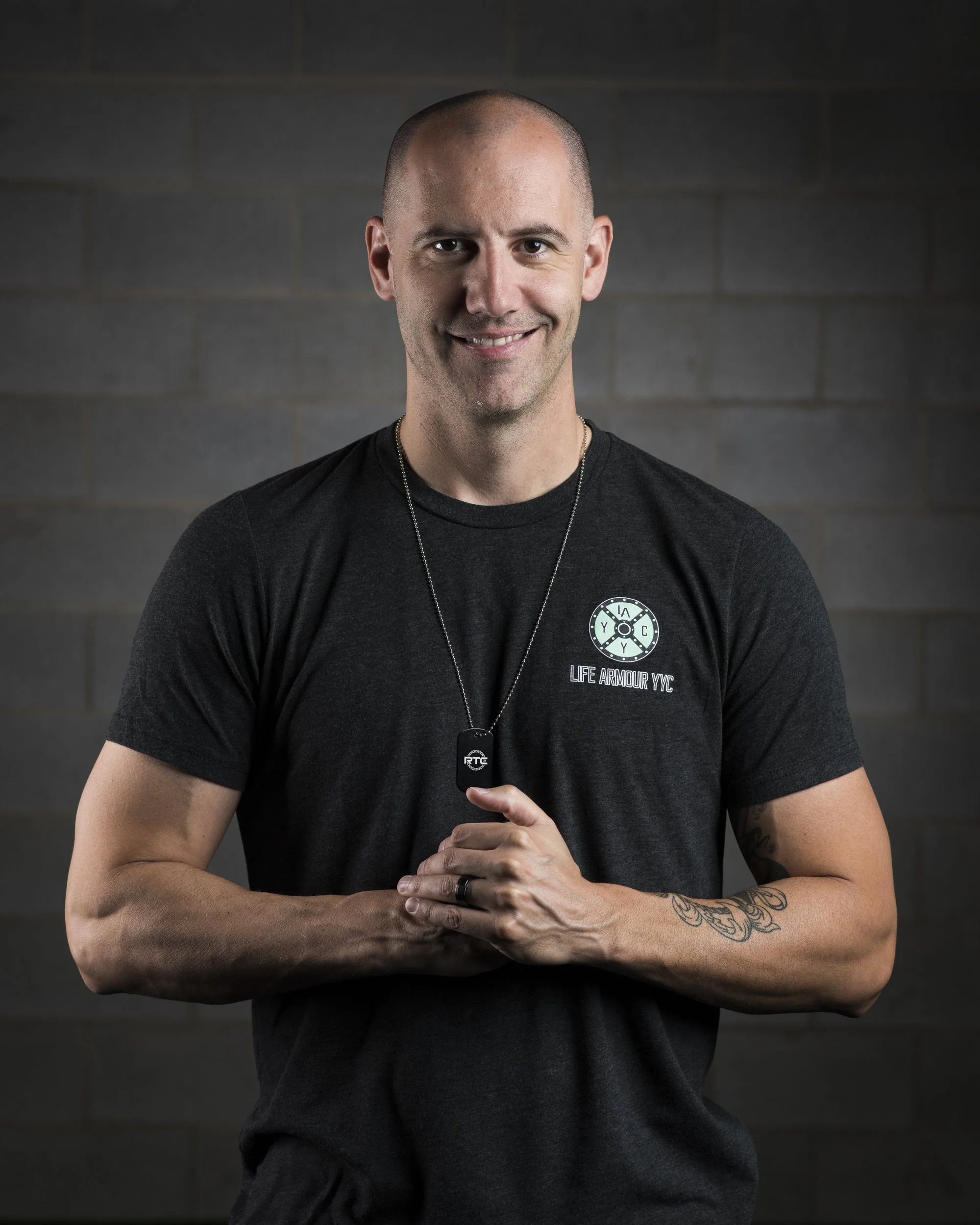 A smiling bald man wearing a black T-shirt with a logo that says 'Life Armour NYC' and a dog tag necklace, standing in front of a dark, brick wall background, with tattoos on his right arm.