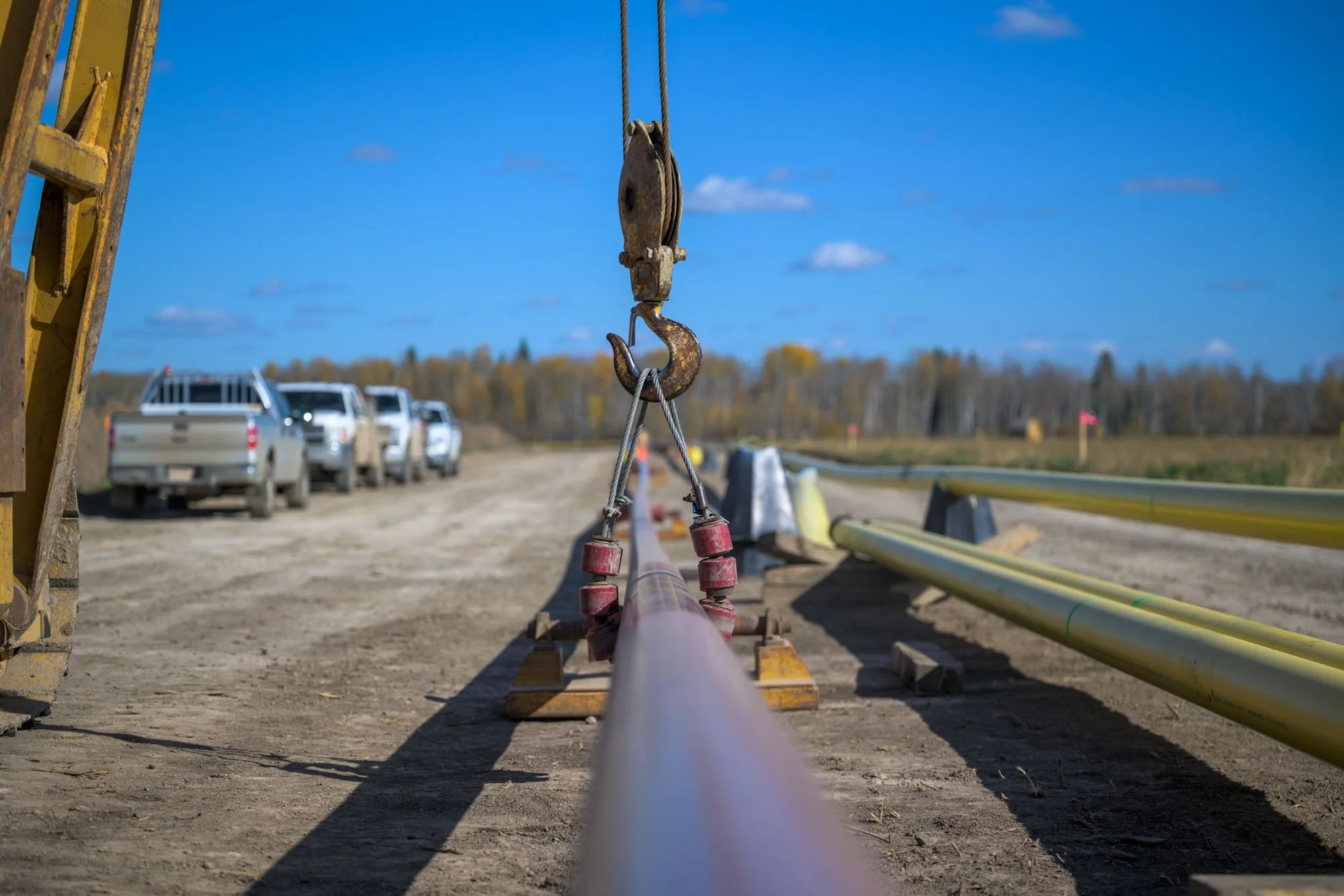 Construction site with yellow pipes lying on the ground, a crane hook holding a pipe, and several trucks in the background under a blue sky.