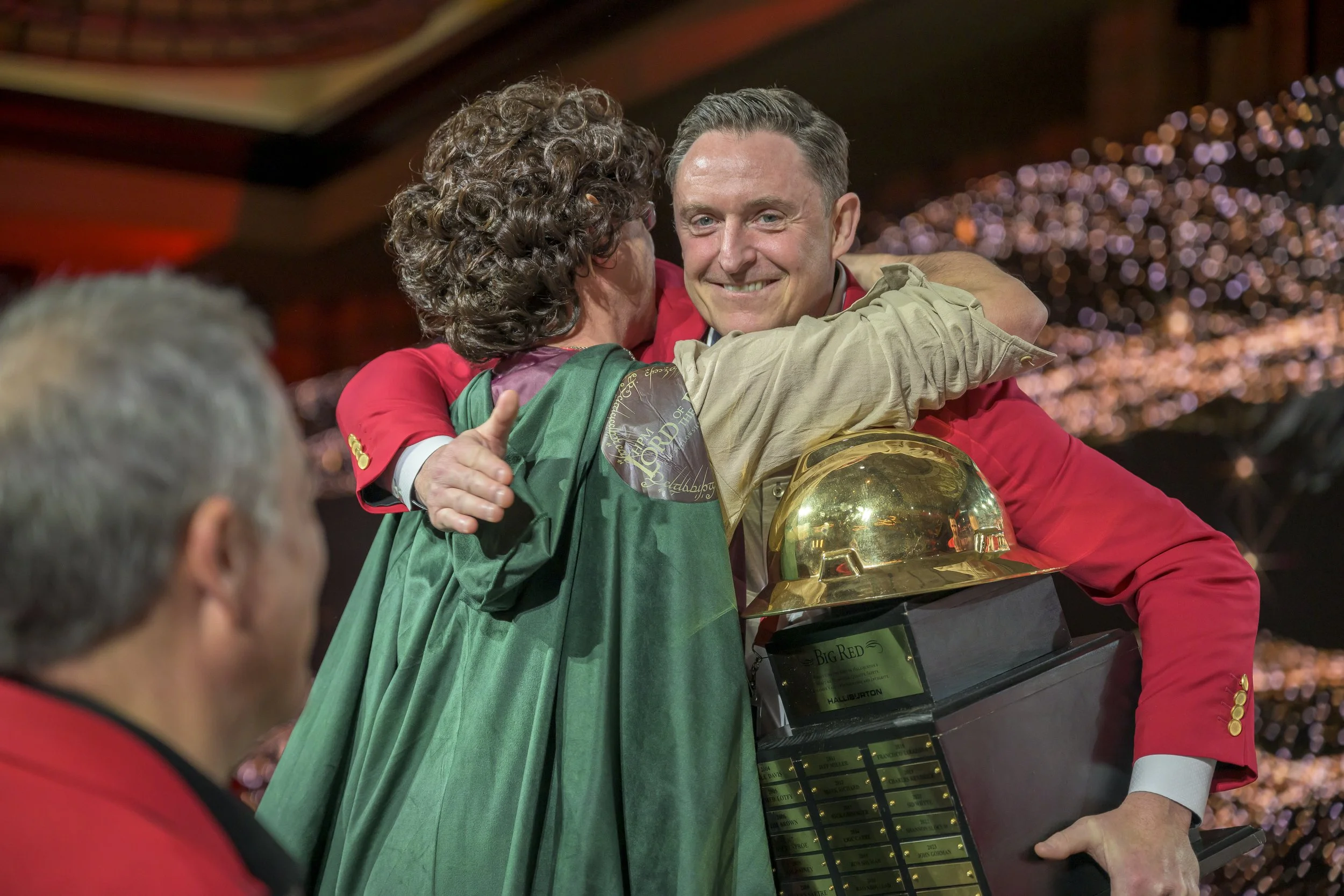 Two men hugging, one man in a red jacket and the other in a costume with a green cape, celebrate on stage with a trophy in a dimly lit room decorated with small string lights.