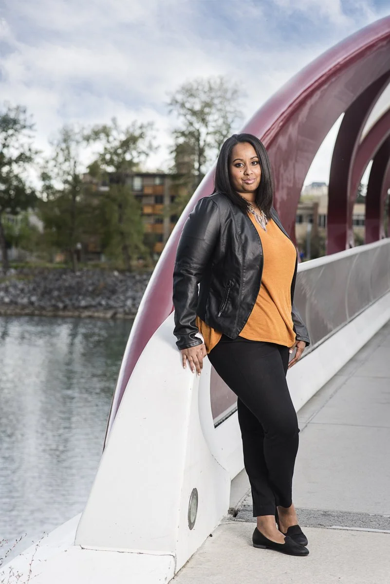 Woman in black leather jacket, orange top, and black pants standing on a bridge with a river and trees in the background.