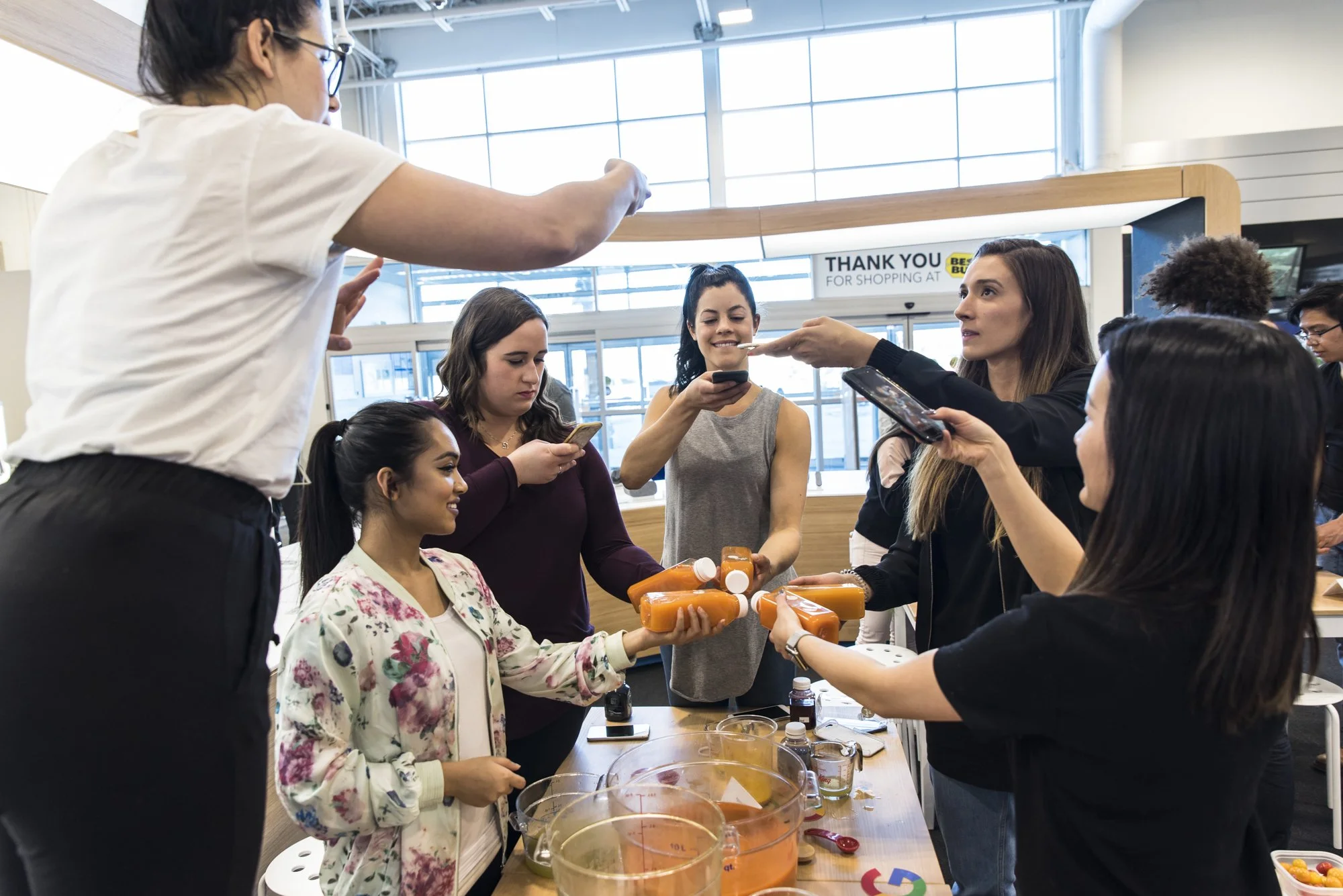 Group of women at a health or wellness event, with one woman handing out bottles and others interacting and taking photos. The setting appears to be an indoor space with large windows.