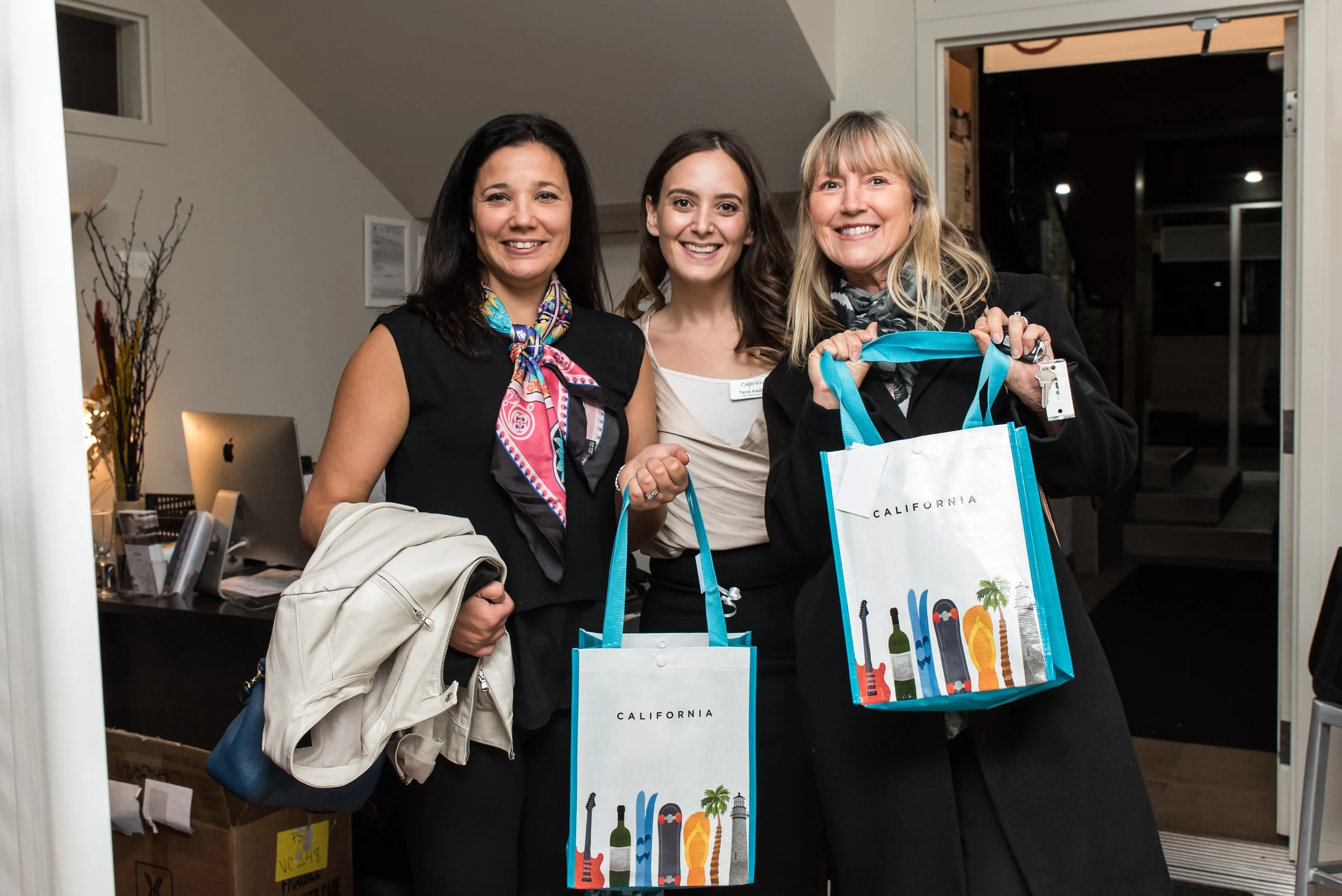 Three women smiling in an office, holding bags with California-themed graphics, posing for a photo.