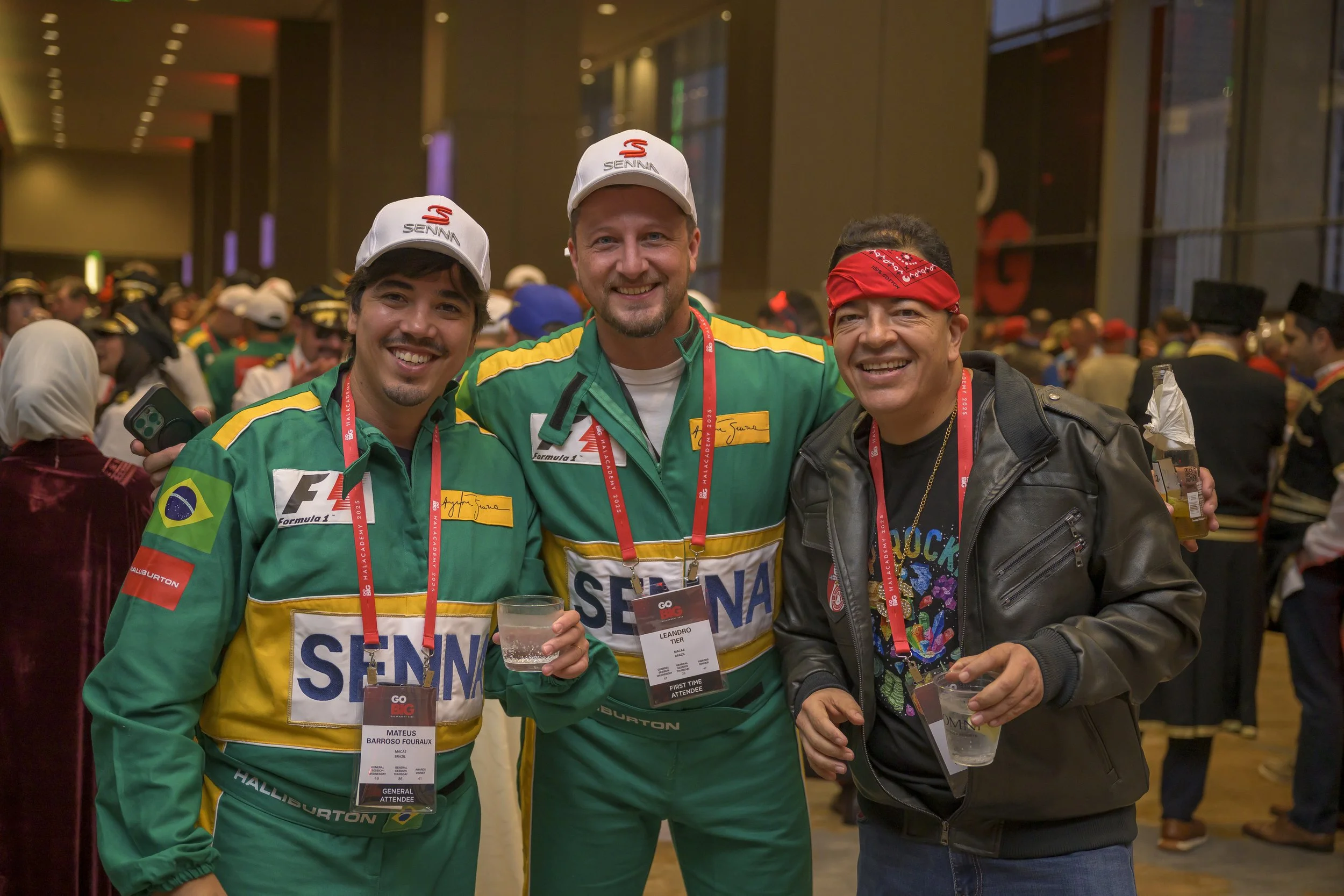 Three men smiling and posing together at a crowded indoor event, with two dressed in green racing uniforms and one in casual clothing with a red bandana and leather jacket, all wearing event badges around their necks, holding drinks.