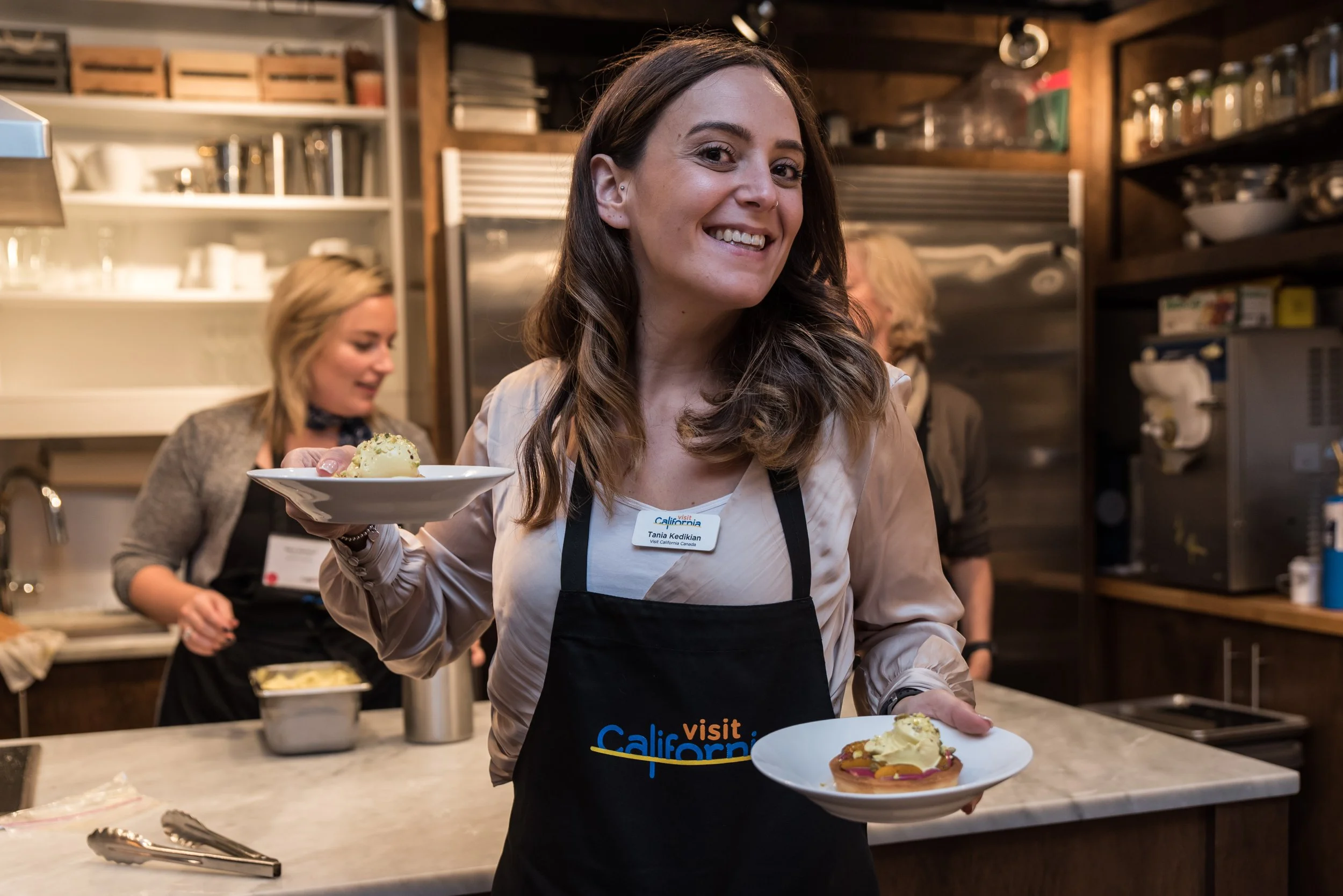 Smiling woman with a name tag and apron holding two plates of dessert in a cozy kitchen with other people in the background.
