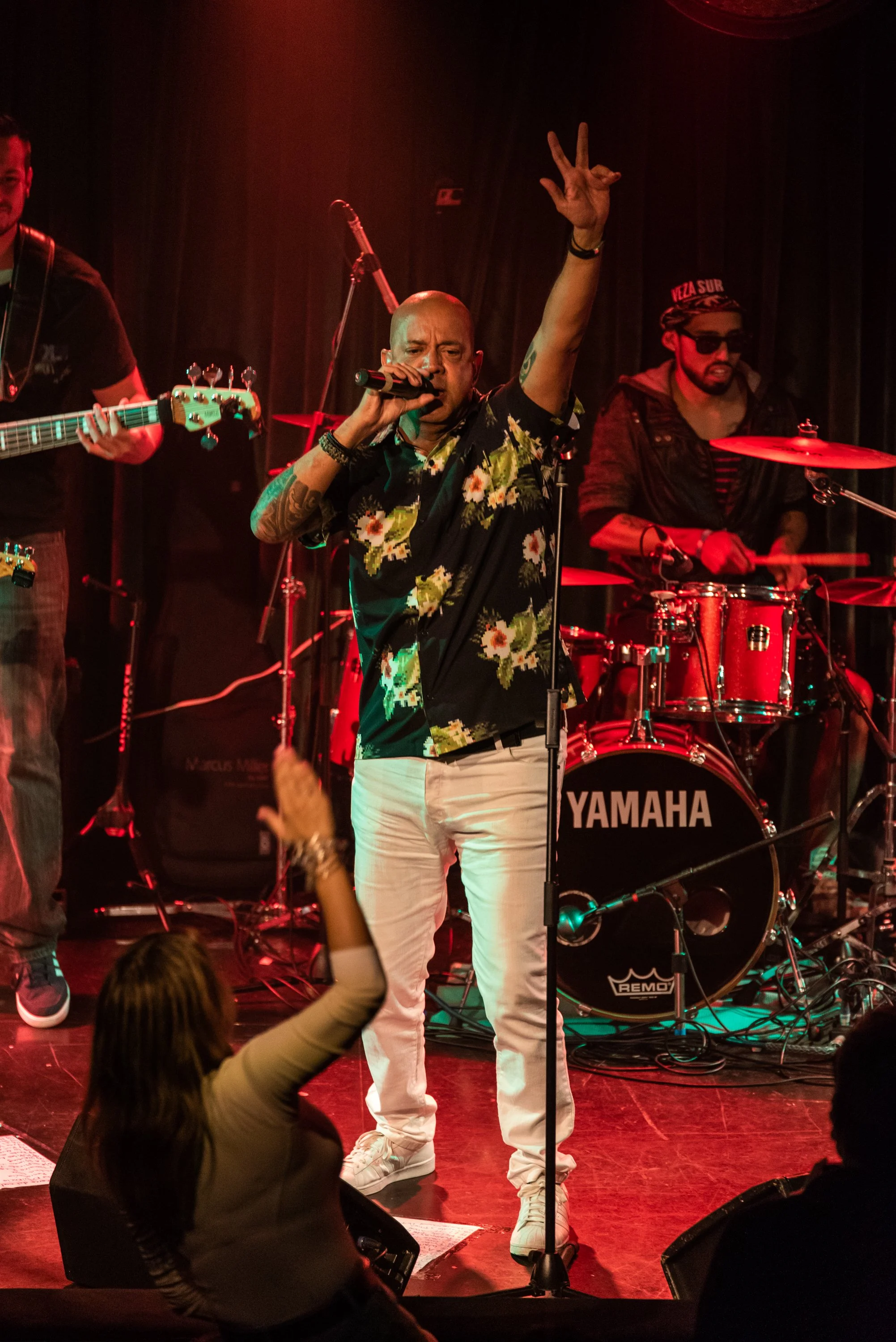 A male singer with a floral shirt and white pants performs on stage with a microphone, raising his right hand with two fingers in a peace sign. Behind him, a drummer with sunglasses and a cap plays a Yamaha drum set. To the left, a guitarist plays an