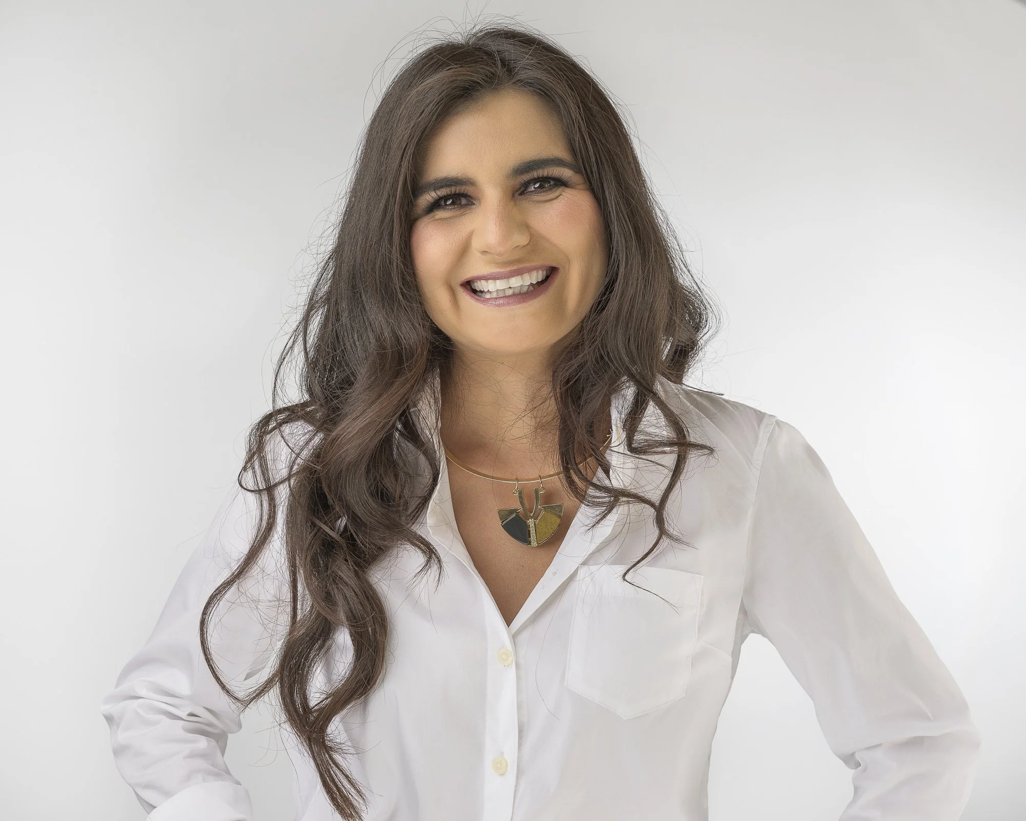 A woman with long wavy brown hair smiling, wearing a white blouse and a gold and black necklace, against a plain white background.