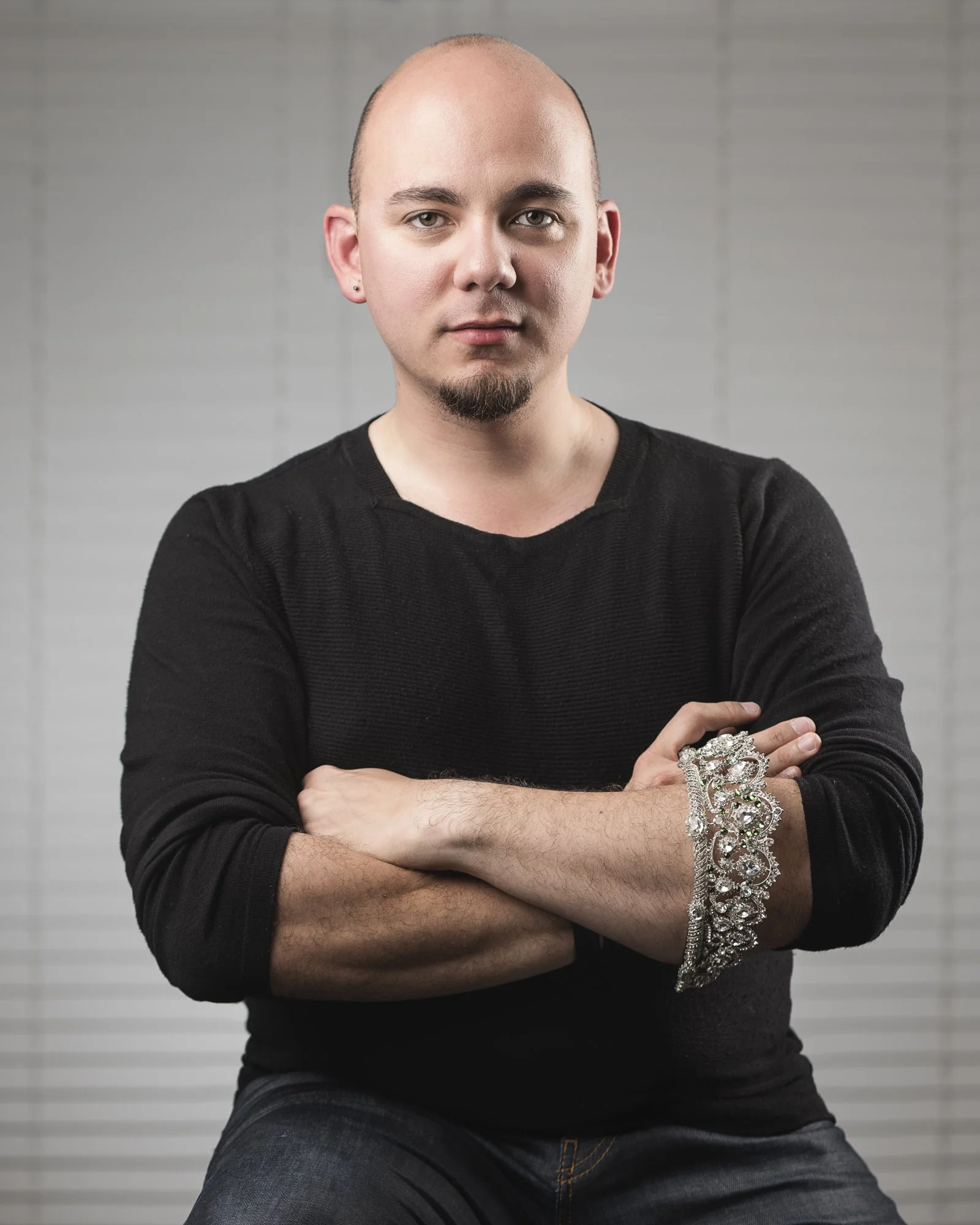 A bald man with a goatee and earrings, wearing a black long-sleeve shirt, sitting with arms crossed, holding a sparkly bracelet on his left arm, against a background of horizontal blinds.