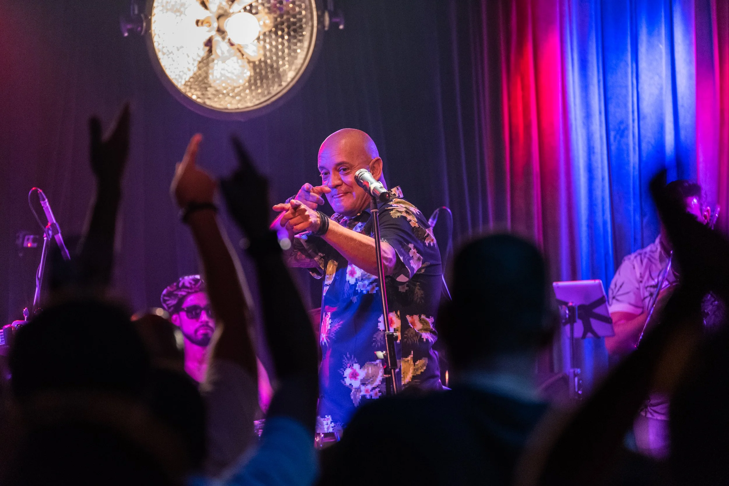 A performer on stage points towards the camera with a smile, wearing a floral shirt, with a microphone in front, and an audience with raised hands in the foreground. The stage has colorful lighting and curtains in the background.