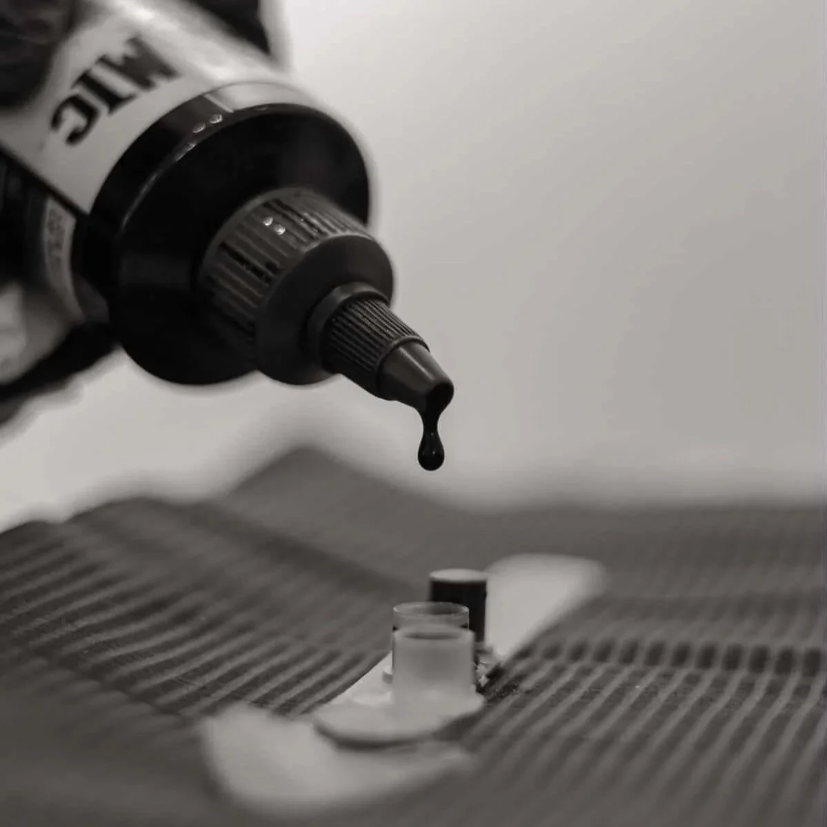 A close-up of a black tube dispensing a dark liquid onto a checkered fabric surface, with a small open container below.