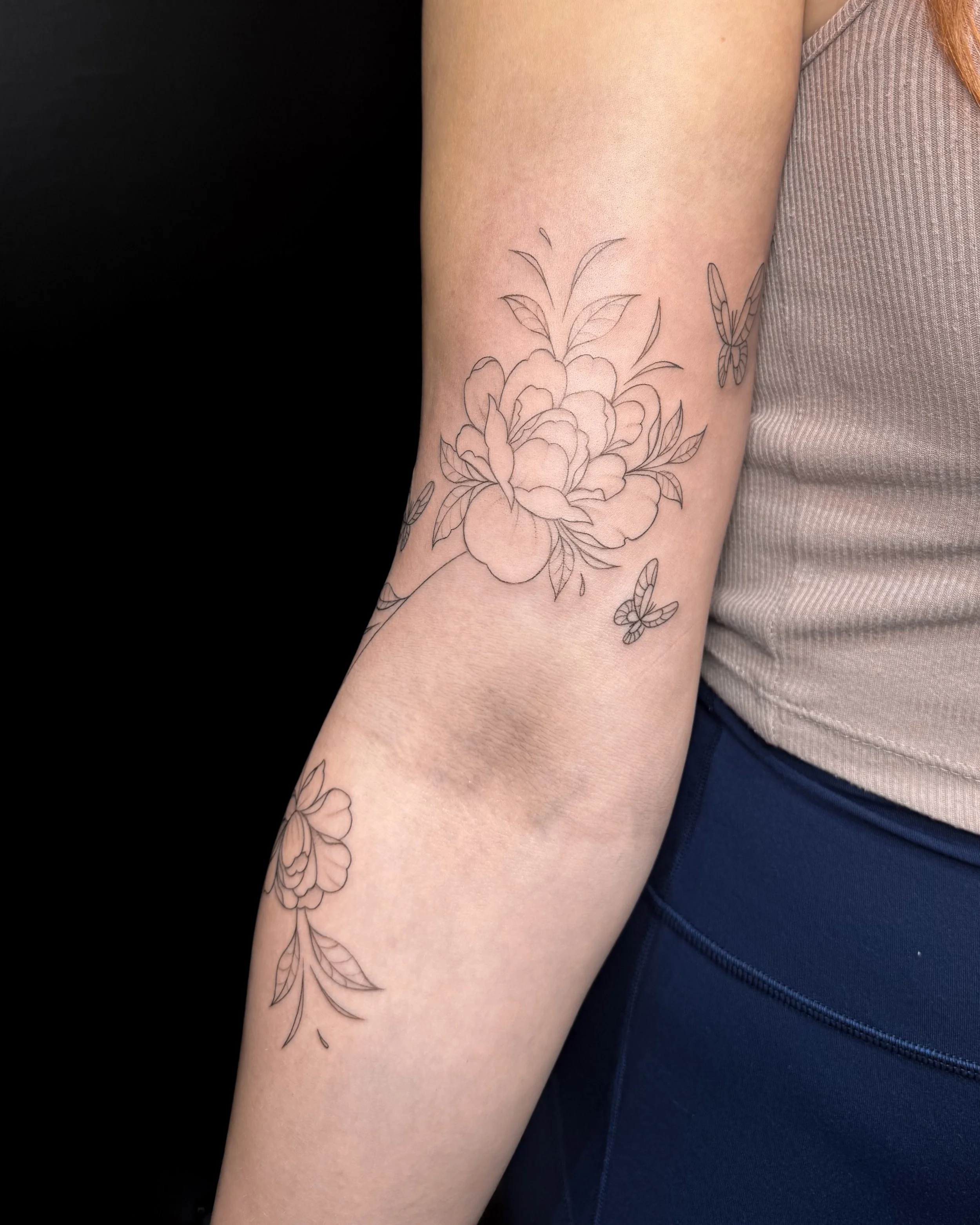 A person's inner arm with a black outline floral tattoo and a small butterfly tattoo nearby, wearing a striped sleeveless top and dark blue pants.