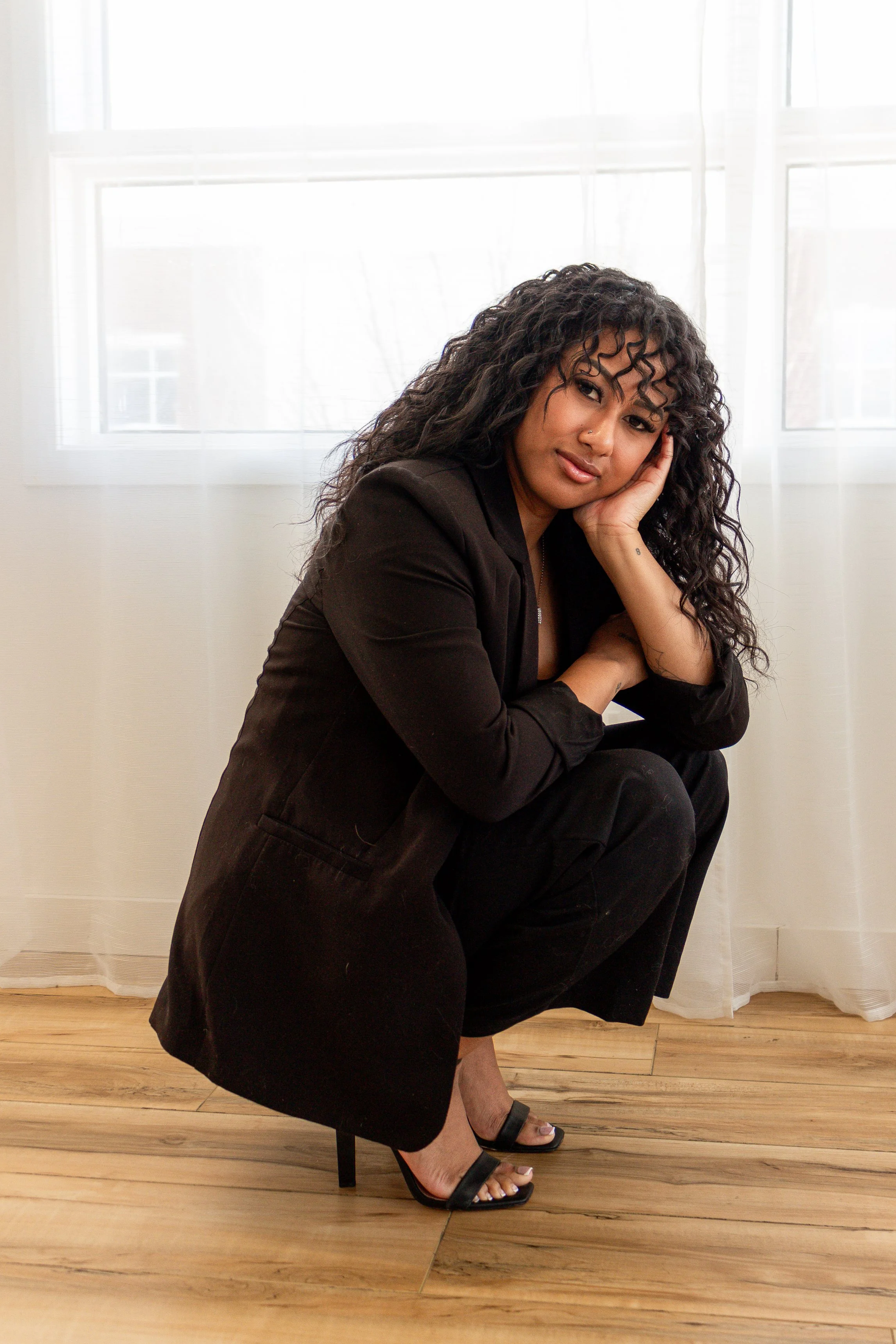 A woman with curly black hair and medium skin tone, wearing a black blazer, black dress, and black high heel sandals, is crouching on a wooden floor in front of a window with white sheer curtains. She is resting her face on her right hand, smiling softly and looking into the camera.