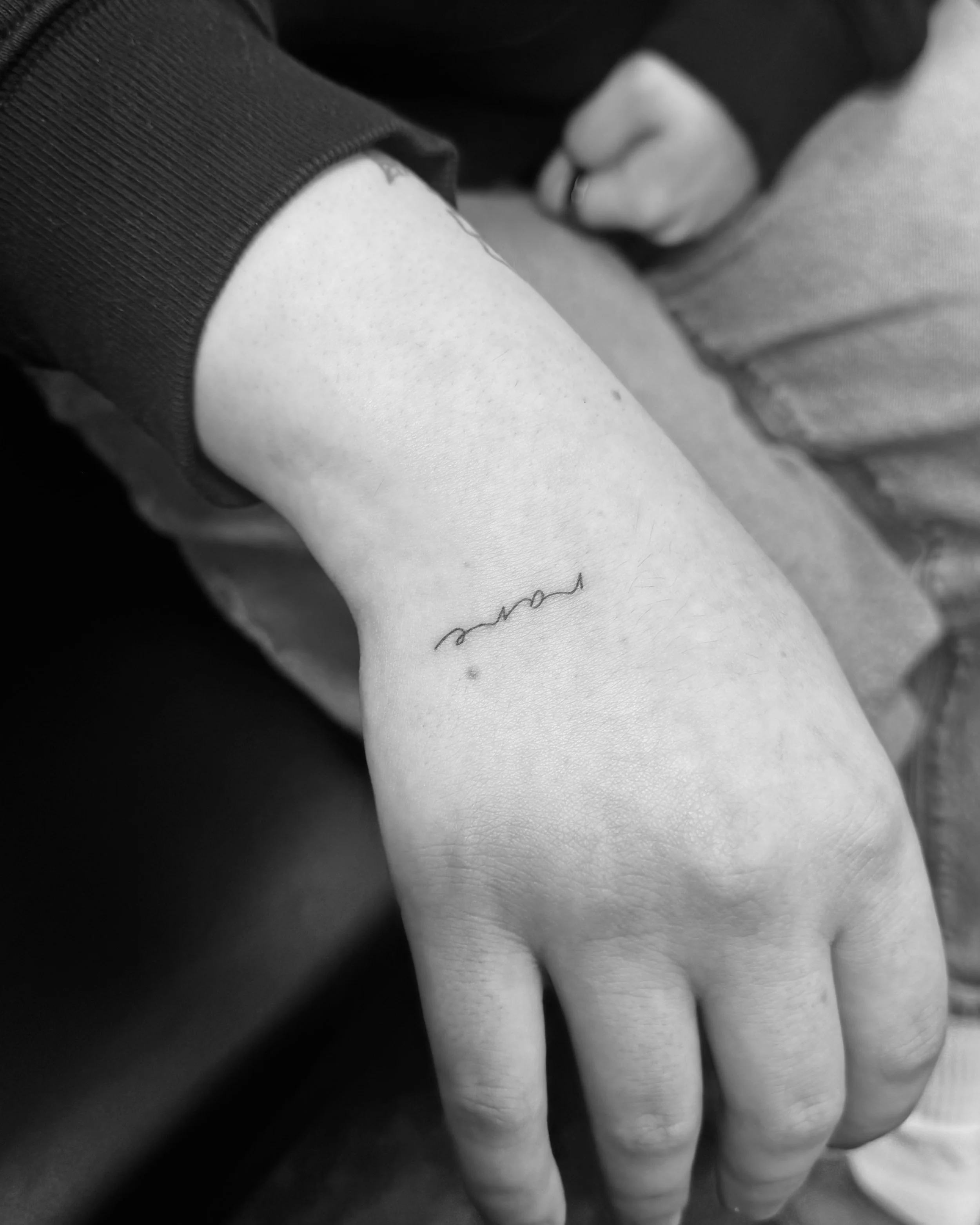 Close-up black and white photograph of a person's hand resting on their thigh, wearing a ring on the middle finger, with visible skin texture and a tattoo.
