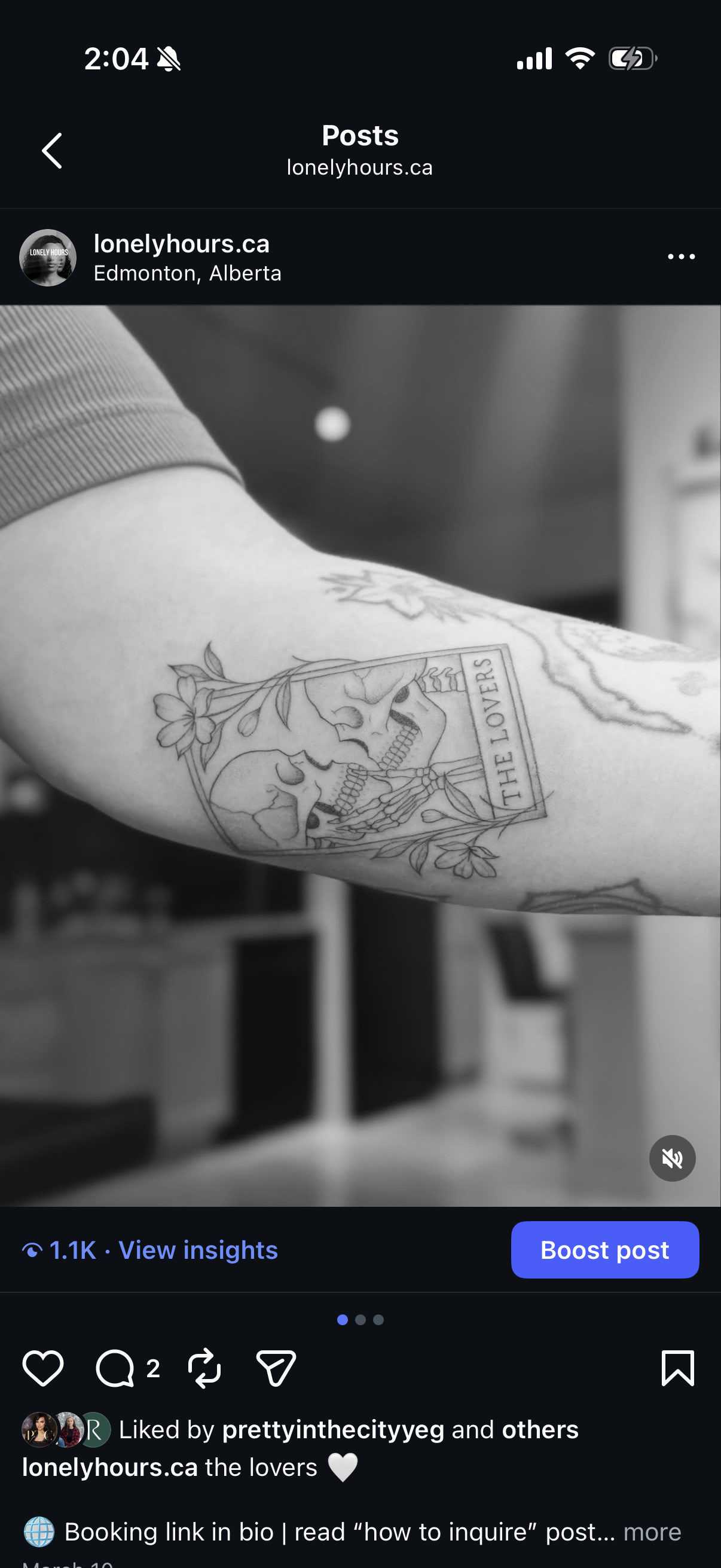 Black and white photo of a person's upper arm with a tattoo of two skeletons, surrounded by flowers, and the text "THE LOVERS" inside a rectangular border.