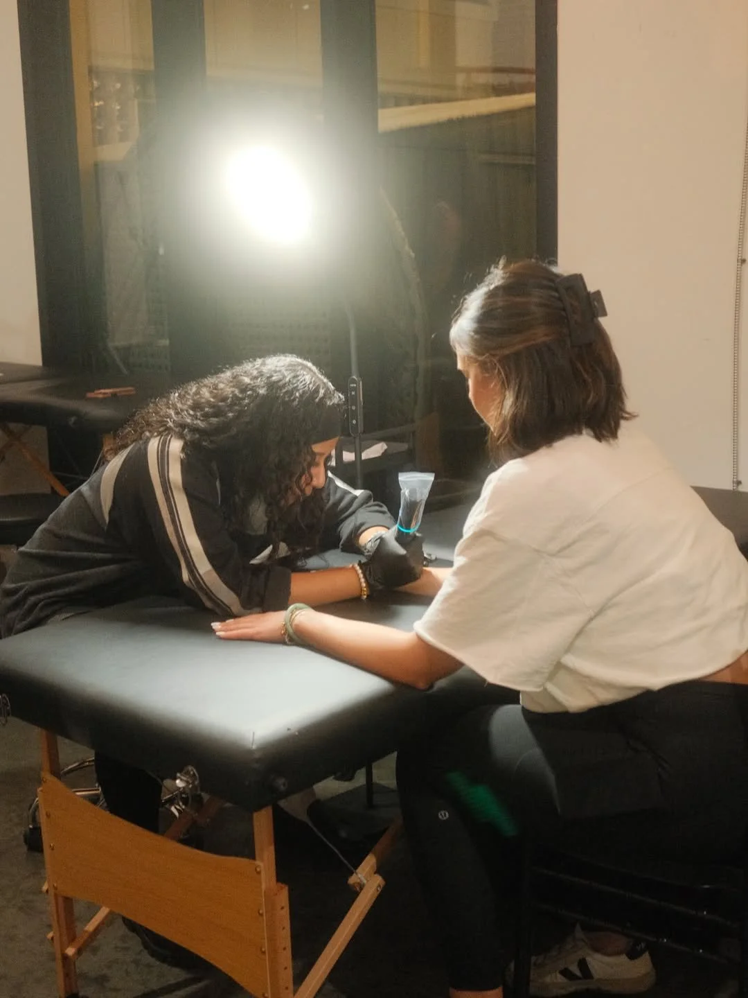 A woman with curly hair lying face down on a padded massage or examination table, receiving a tattoo or similar procedure from another woman with straight hair, seated beside the table. There is a bright light source and some equipment in the background.