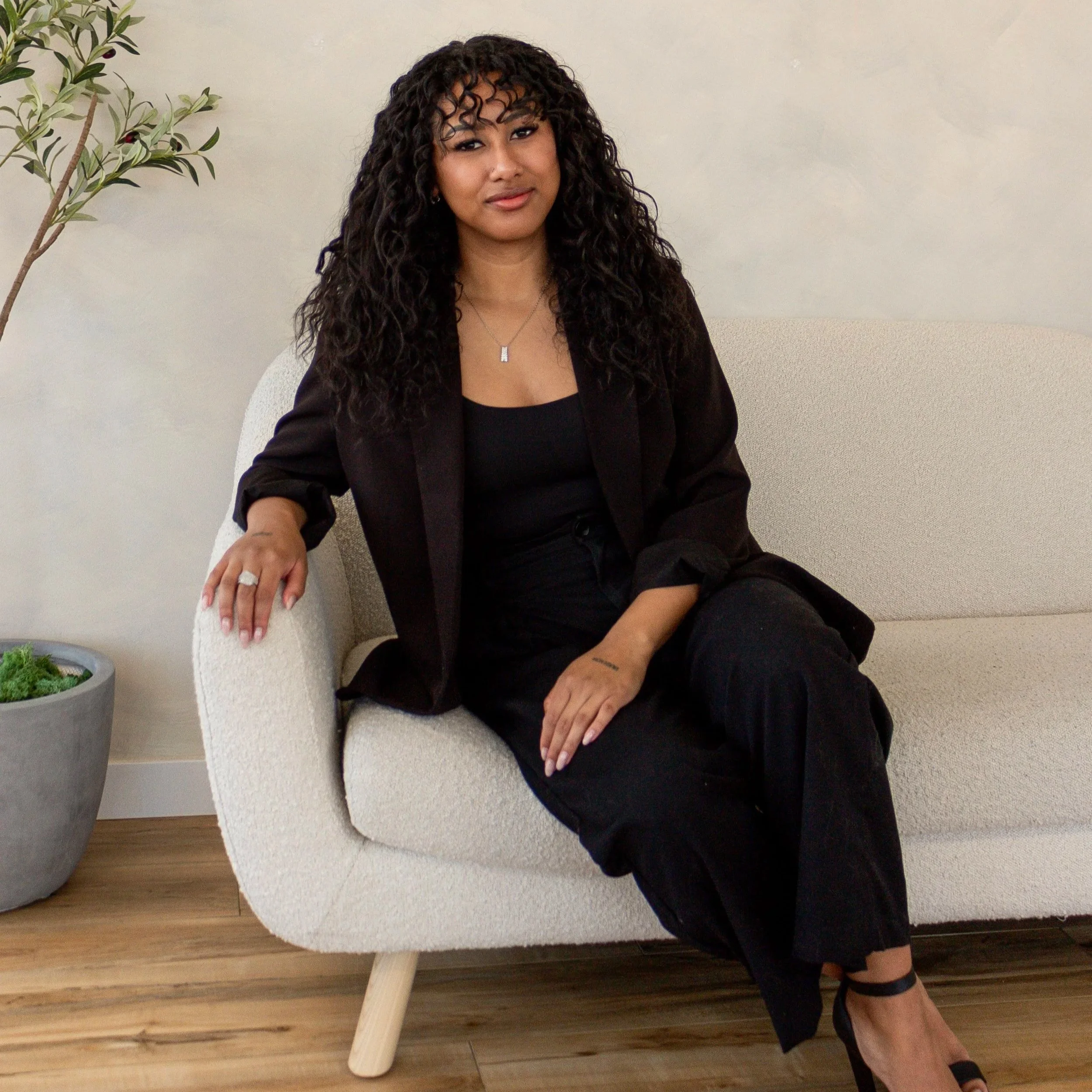 A woman sitting on a white curved chair with a plant nearby, wearing a black outfit and black shoes, with curly hair and a small necklace.