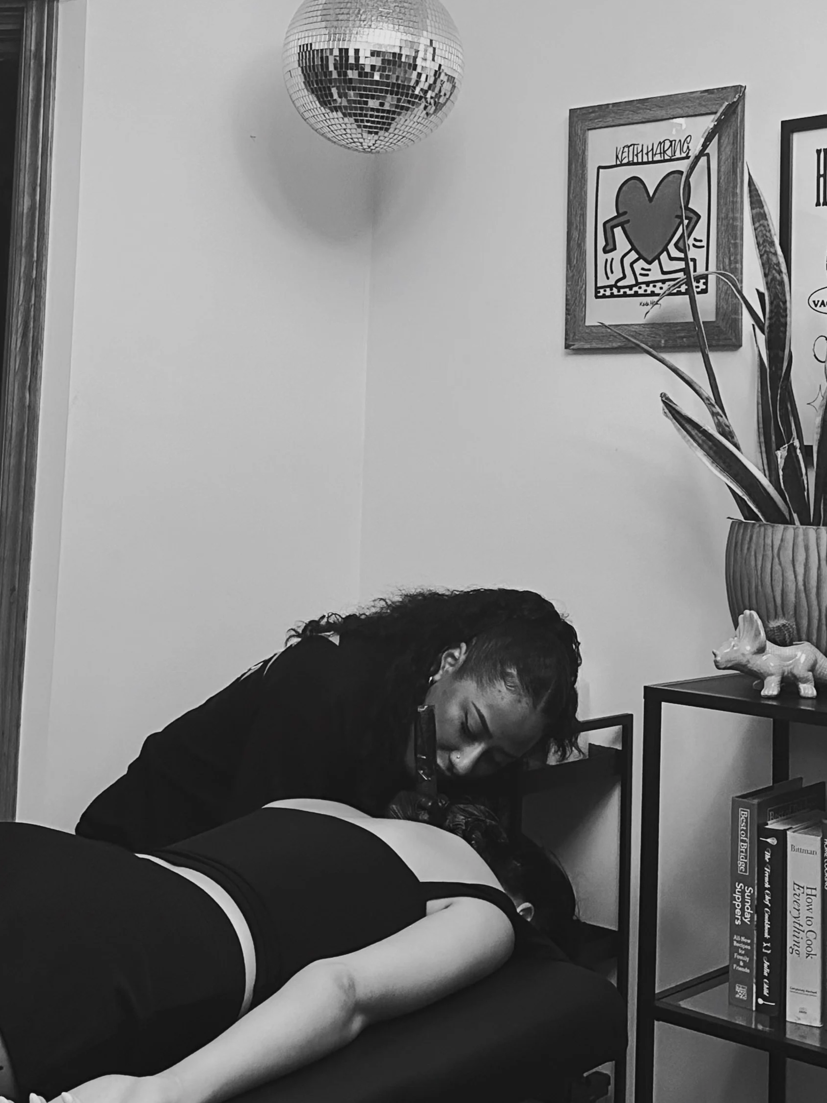 A person with curly hair lying on a couch or bed with their arm extended, wearing a sleeveless top. Another person with straight hair is sitting beside them, resting their head on the first person's arm, in a cozy indoor setting with a wall, a framed artwork, a plant, and a bookshelf that has books and a decorative ceramic dog.