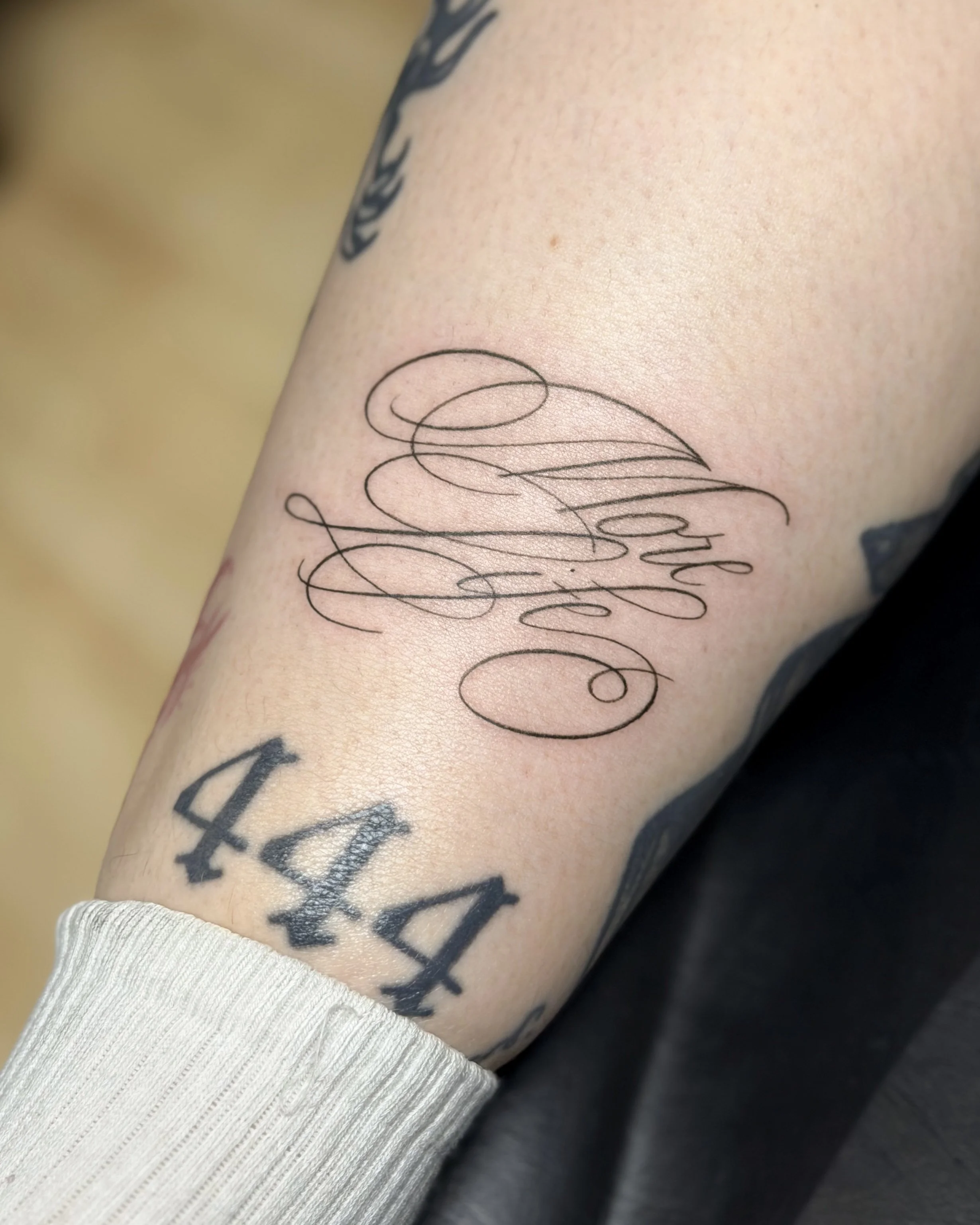 Close-up of a person's forearm with black tattooed text and a delicate line drawing tattoo, wearing a white sleeve at the wrist.