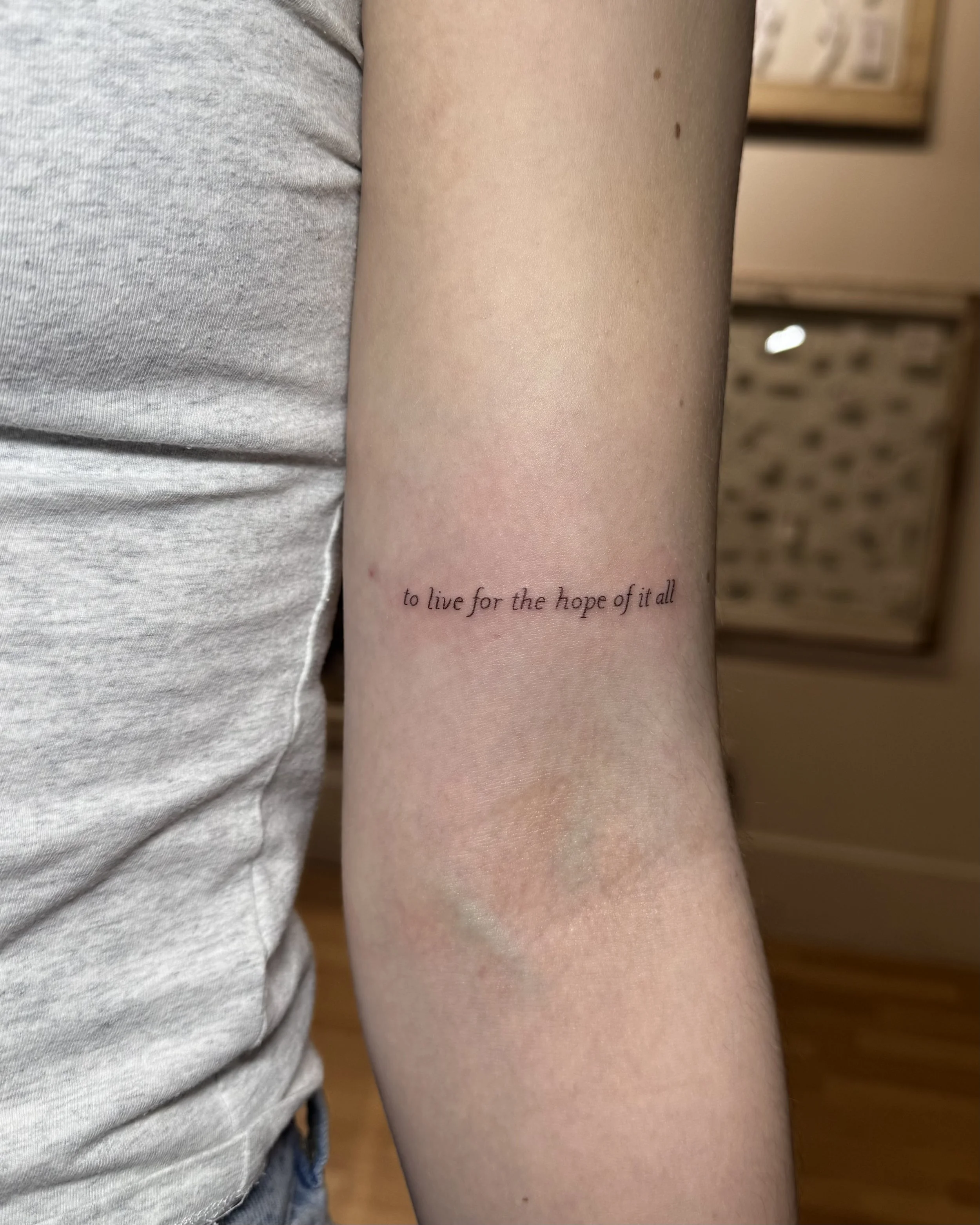 Close-up of a person's arm with a tattoo reading 'to live for the hope of it all' in small, black cursive font.
