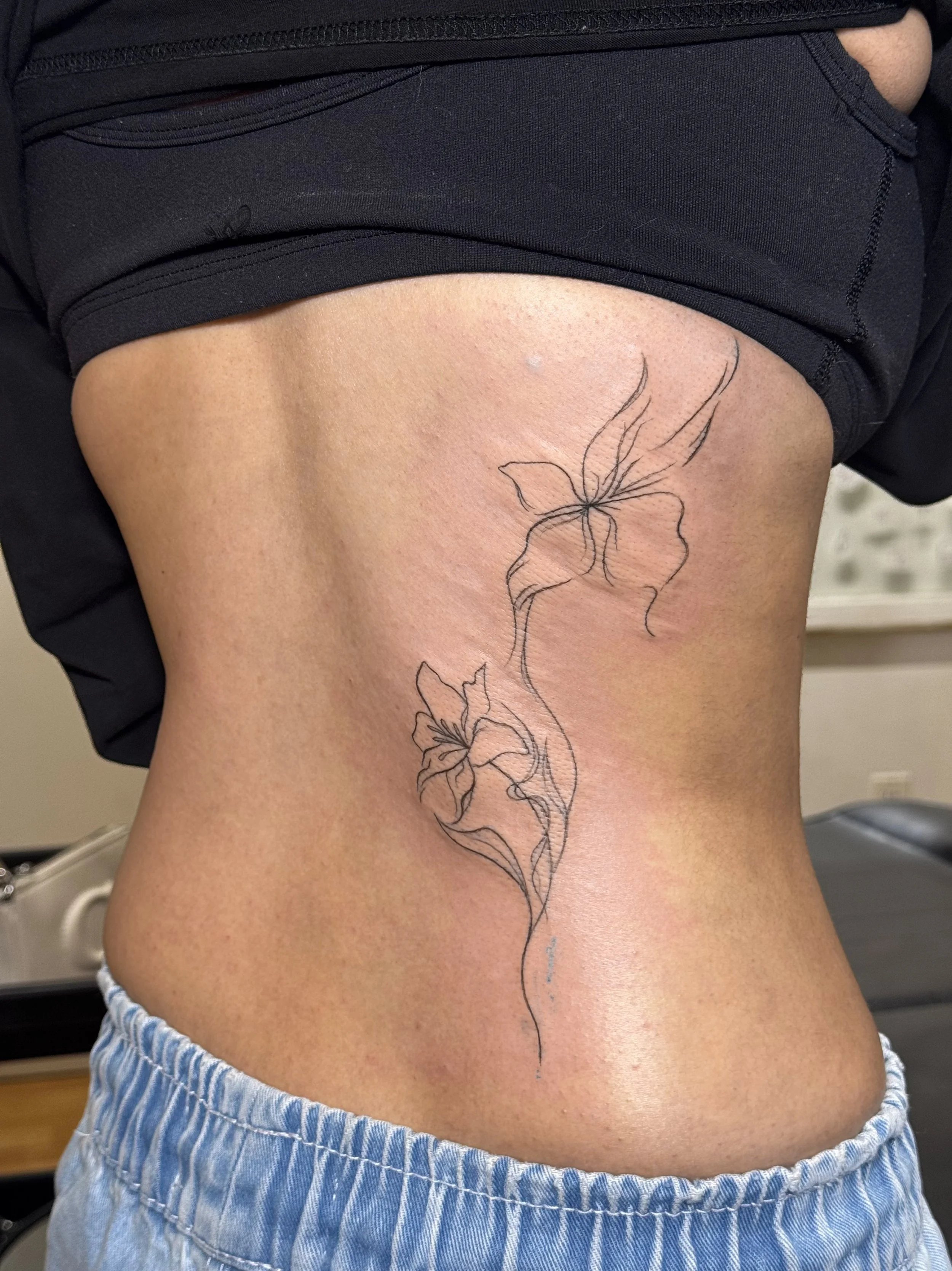 A person's side torso with a black outline tattoo of two butterflies connected by a vine, wearing a black shirt and light blue and white striped shorts.