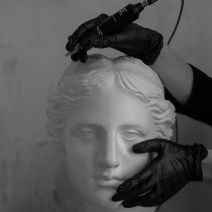 Person's hands, wearing black gloves, are working on a classical-style stone bust sculpture with a drill or rotary tool.