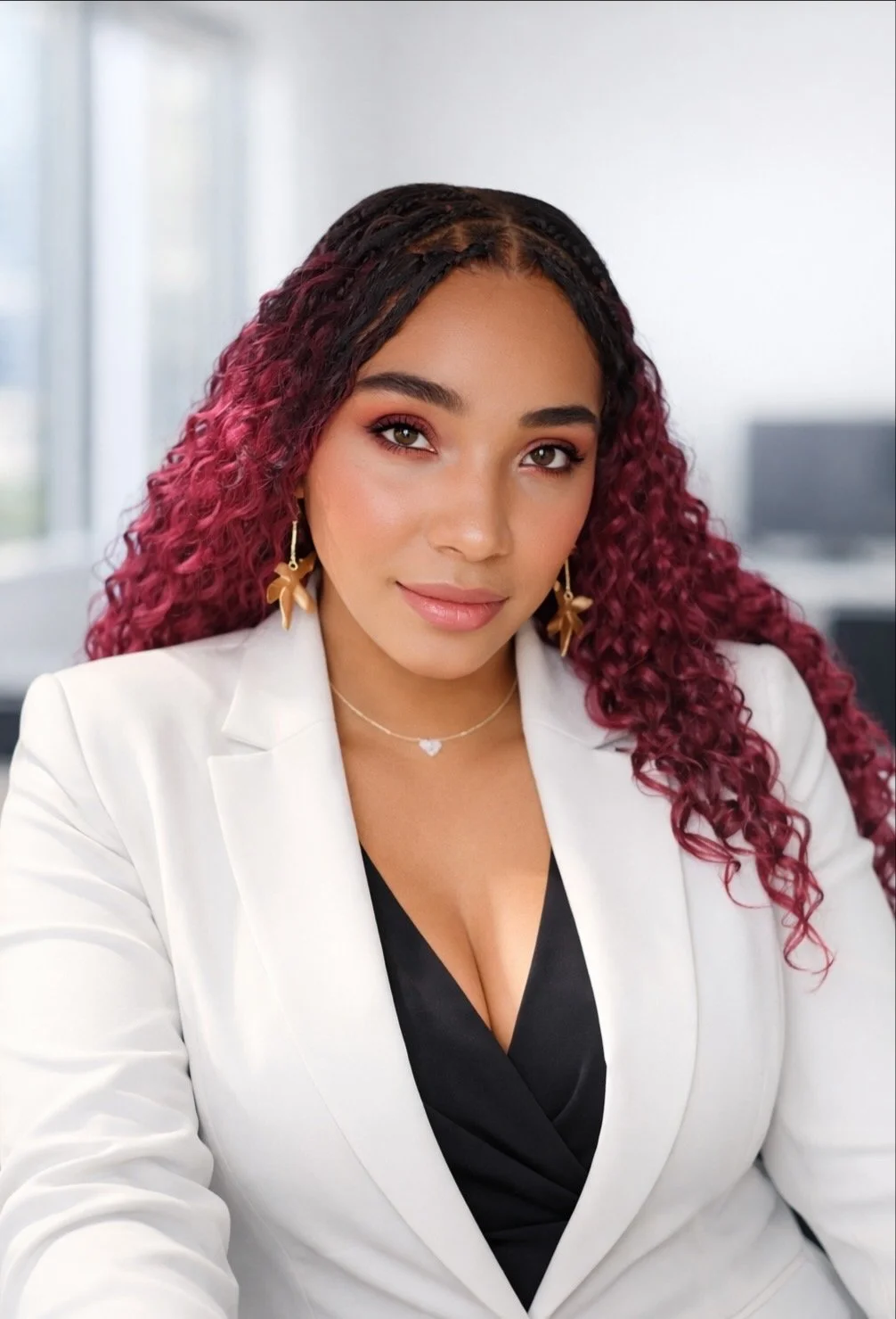 A woman with curly reddish hair and black hair, wearing a white blazer and black top, sitting in an office setting with large windows in the background.