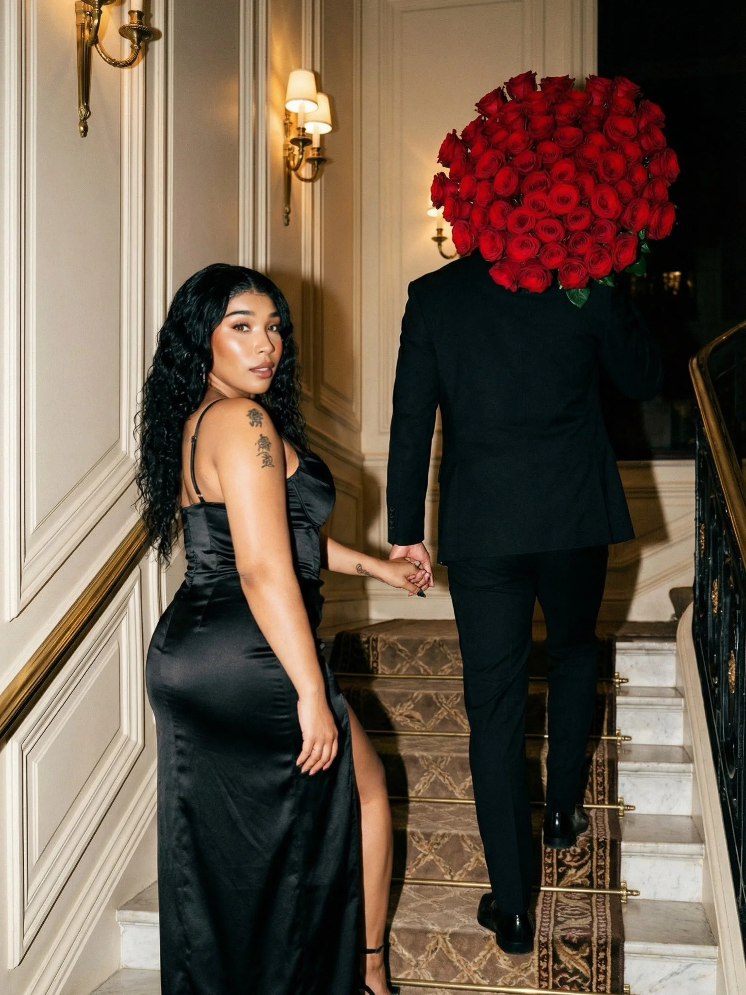 A woman in a black satin dress holding hands with a man dressed in a black suit. The man has a large bouquet of red roses covering his head. They are ascending a staircase inside an elegant, well-lit room.