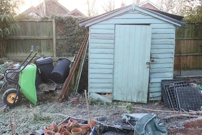 A blue wooden shed in a backyard, surrounded by gardening tools, plant pots, and miscellaneous outdoor items.