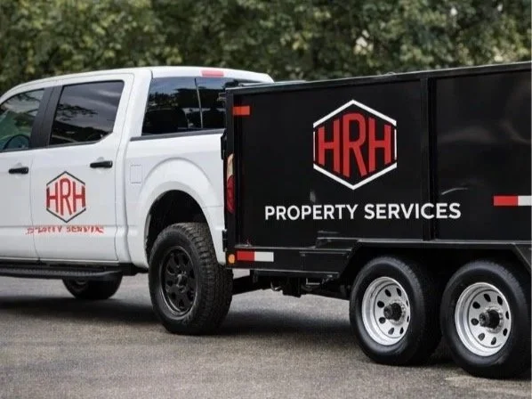 A white pickup truck and a black trailer with HRH Property Services logo parked outdoors near trees.