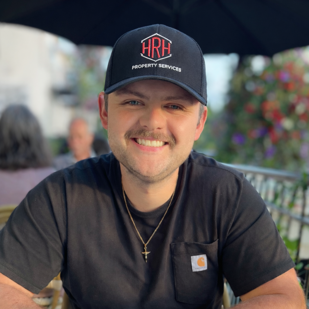 A smiling man wearing a black Carhartt t-shirt and a black baseball cap with the logo 'HRH Property Services'. He has a light beard and mustache, a gold chain with a cross pendant, and is sitting outdoors with a background of colorful flowers and blurred people.