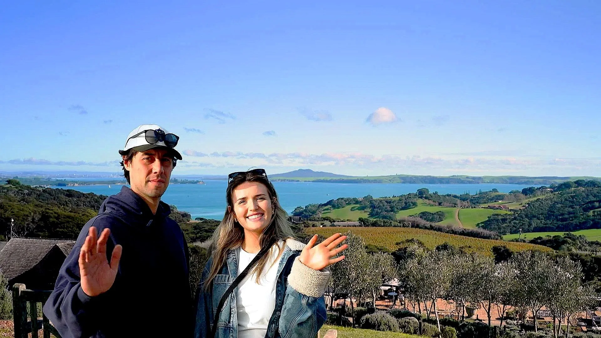 A man and woman standing outdoors near a scenic coastal landscape, smiling and waving at the camera with water and green rolling hills in the background.
