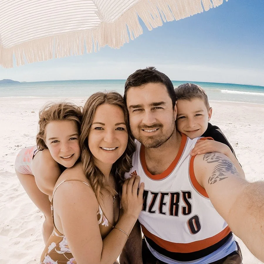 A family of four taking a selfie on the beach under a pink and white beach umbrella. The ocean with waves and a distant coastline can be seen in the background.