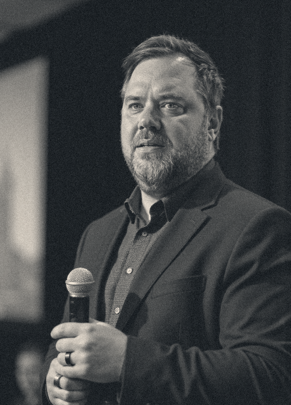 A man with a beard and short hair holding a microphone, dressed in a dark suit jacket and shirt, appearing to speak or present.