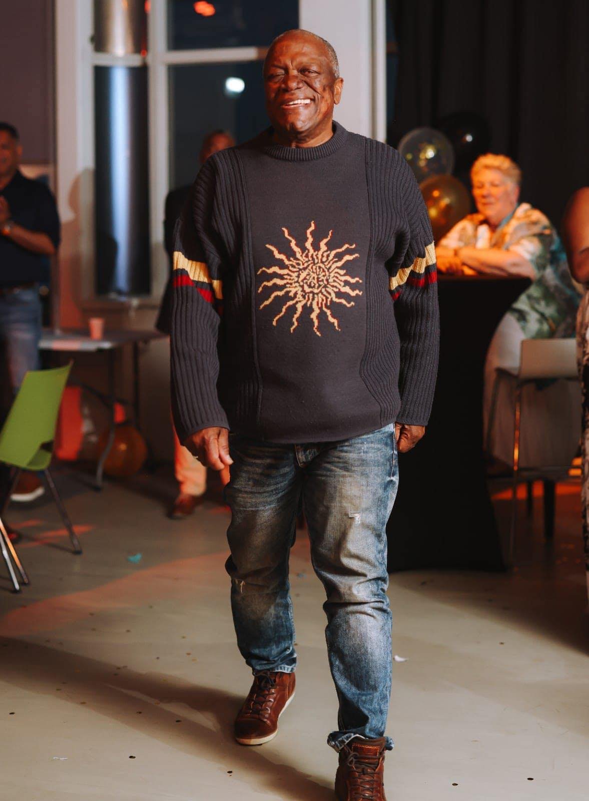 A smiling man walking in a celebration or party setting, wearing a black sweater with gold and red stripe details on the sleeves and a sunburst pattern on the front, paired with distressed jeans and brown boots. In the background, other people are present, some sitting and some standing, in a brightly lit indoor space.