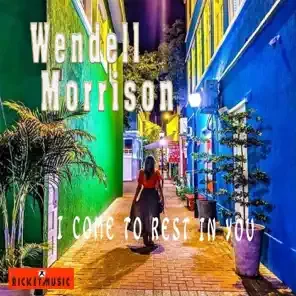 A woman walking through a colorful outdoor corridor with vibrant walls in yellow, blue, and green, featuring tall potted plants and street lamps, with text overlay mentioning Wendell Morrison and 'Come to Rest in Peace,' along with a small logo at the bottom left.