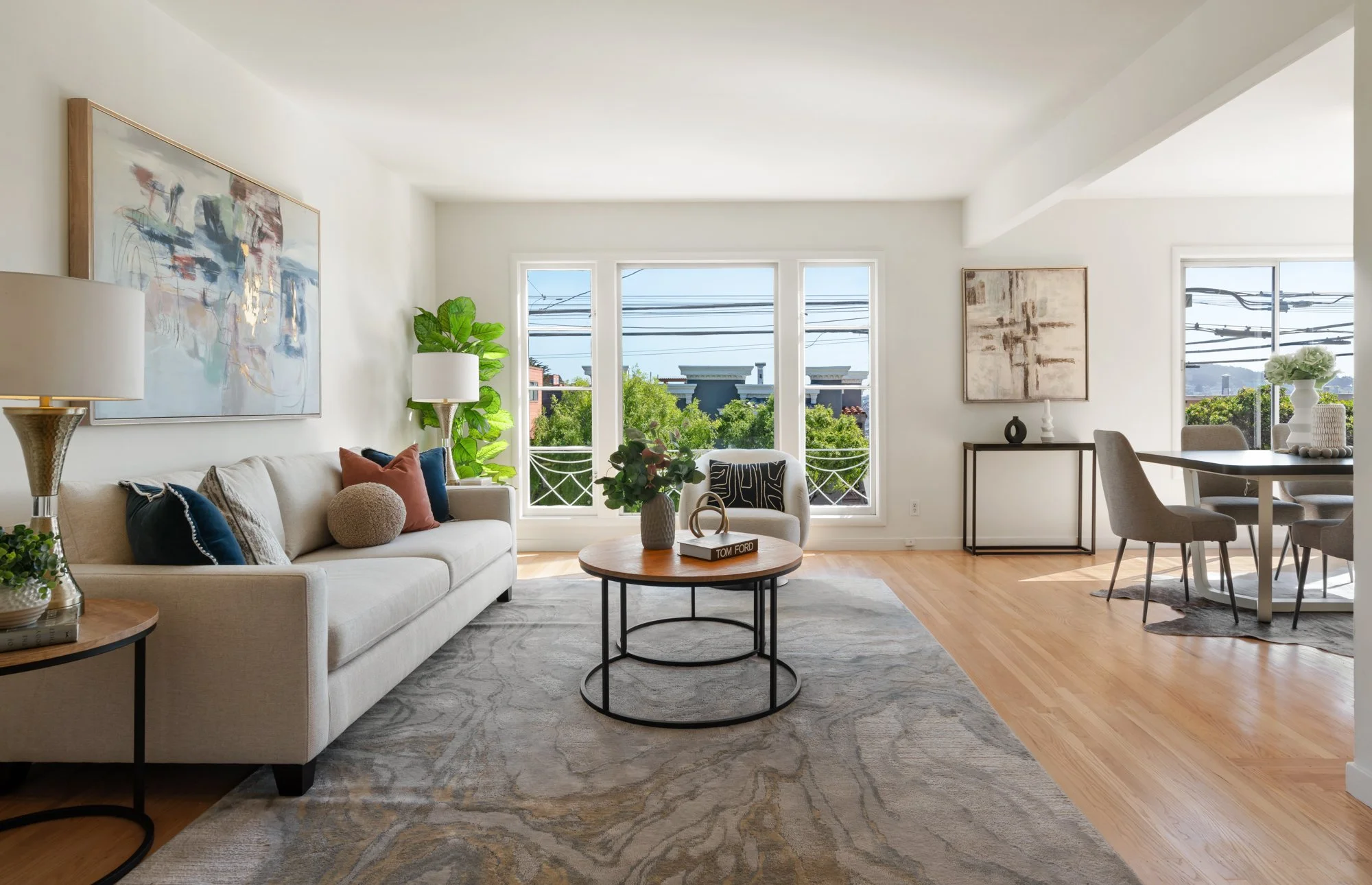 Bright living room with beige sofa, round wooden coffee table, gray rug, white walls, large windows with trees outside, dining area with gray chairs, modern artwork, and houseplants.