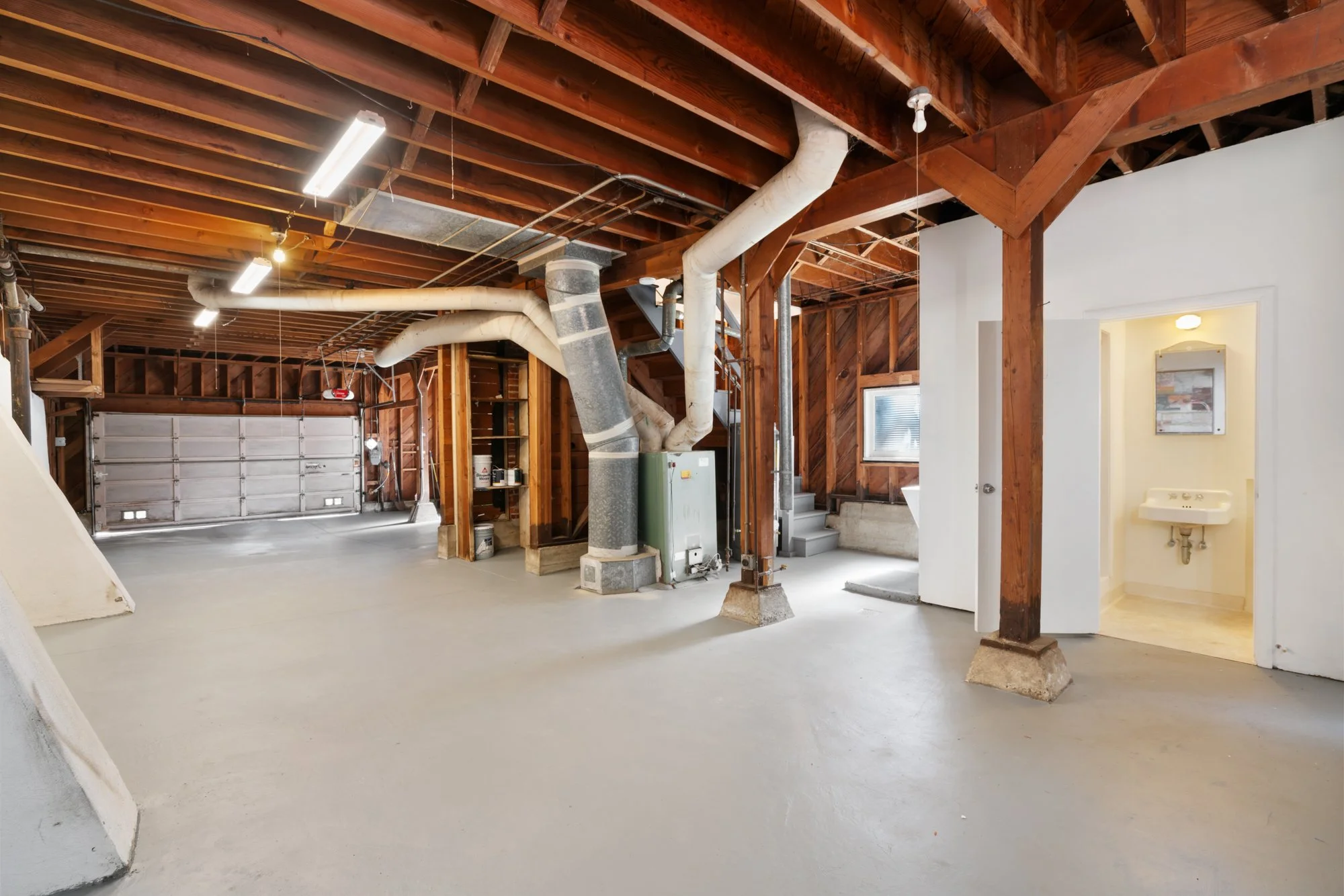 Empty garage with exposed wooden beams, ductwork, and a closed garage door in the background. Small utility room visible to the right.